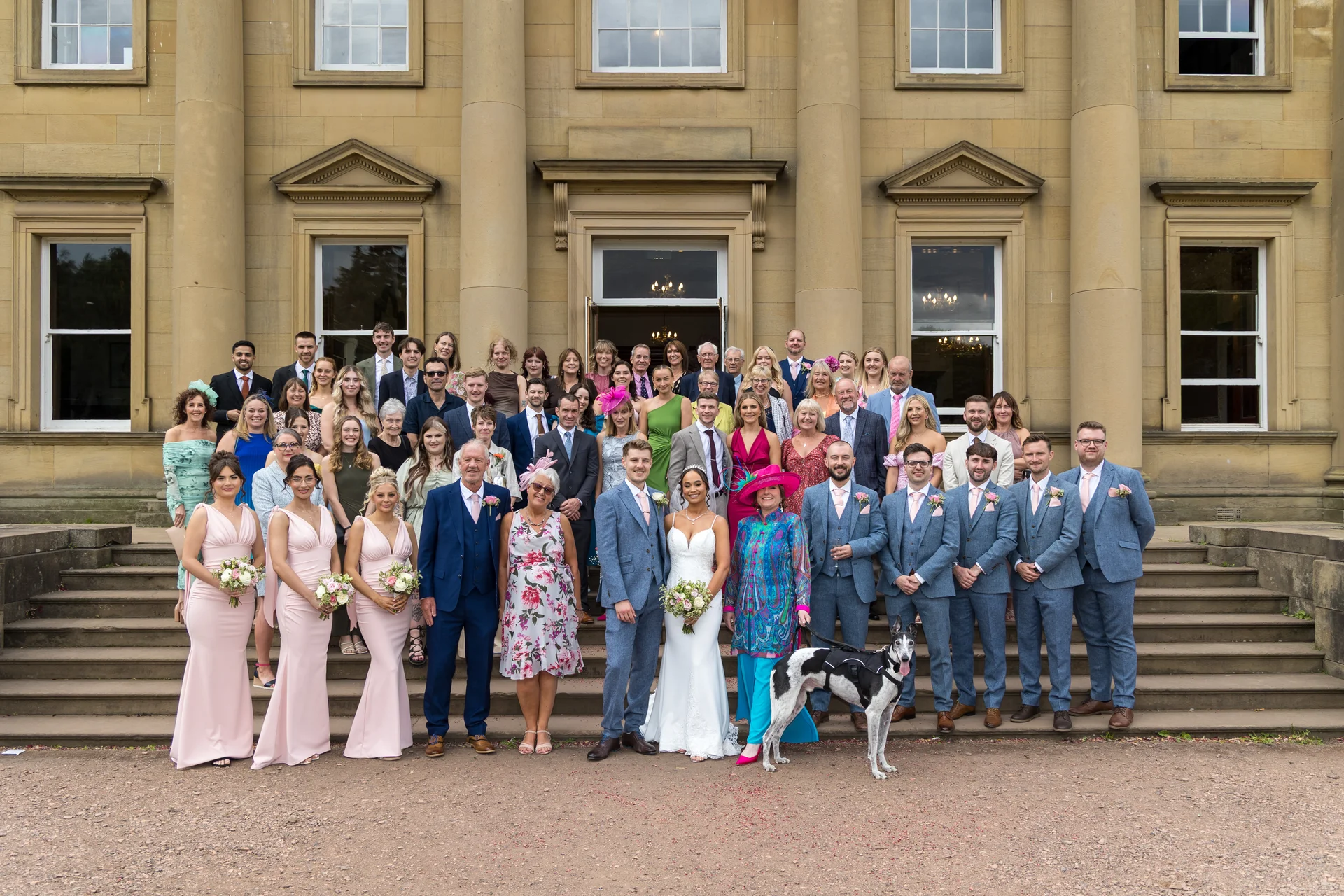 A large wedding party and guests pose for a group photo outside the grand entrance of a stately stone country house.