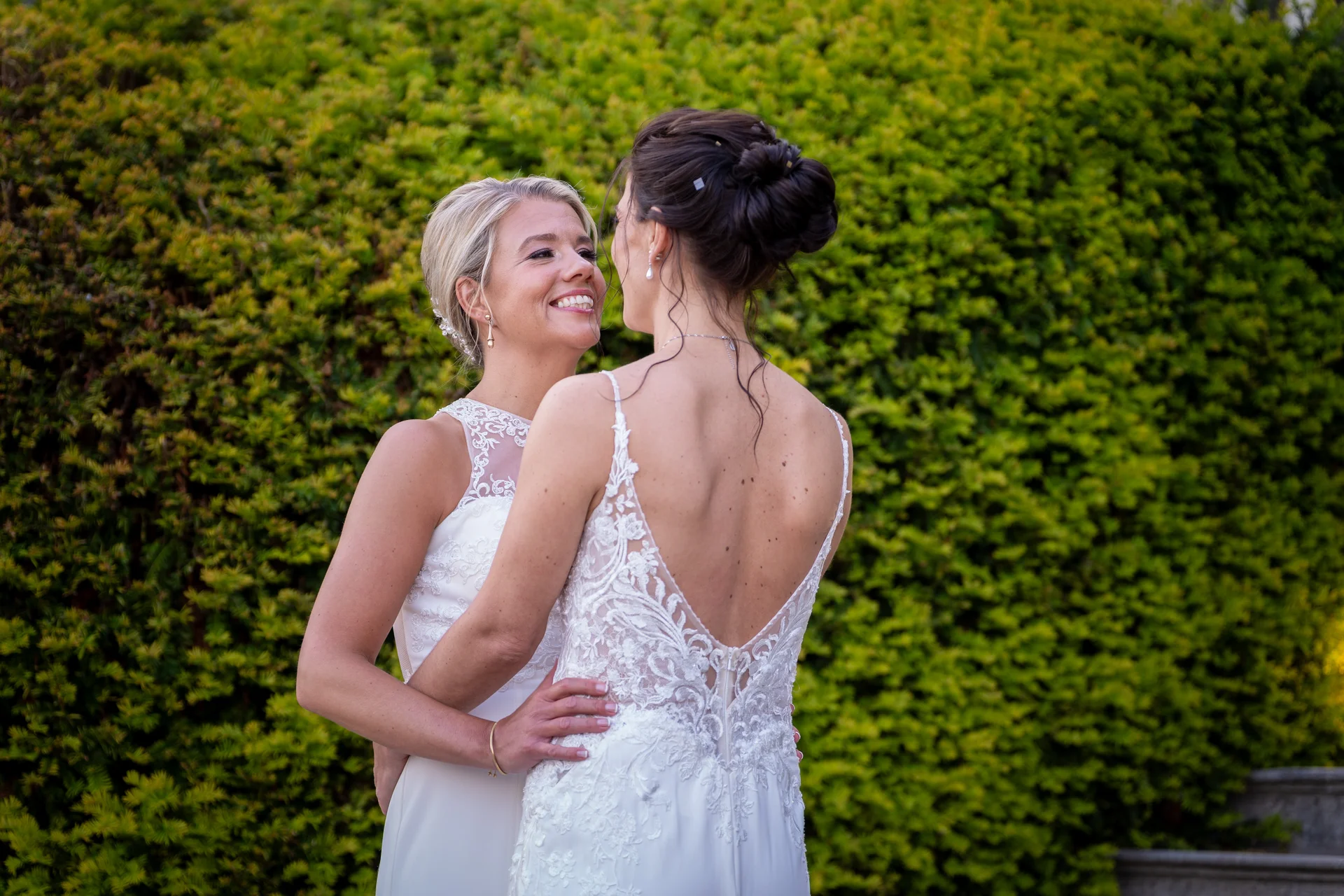 Two smiling brides in white lace wedding gowns embrace warmly against a lush green hedge backdrop during their outdoor portrait session.
