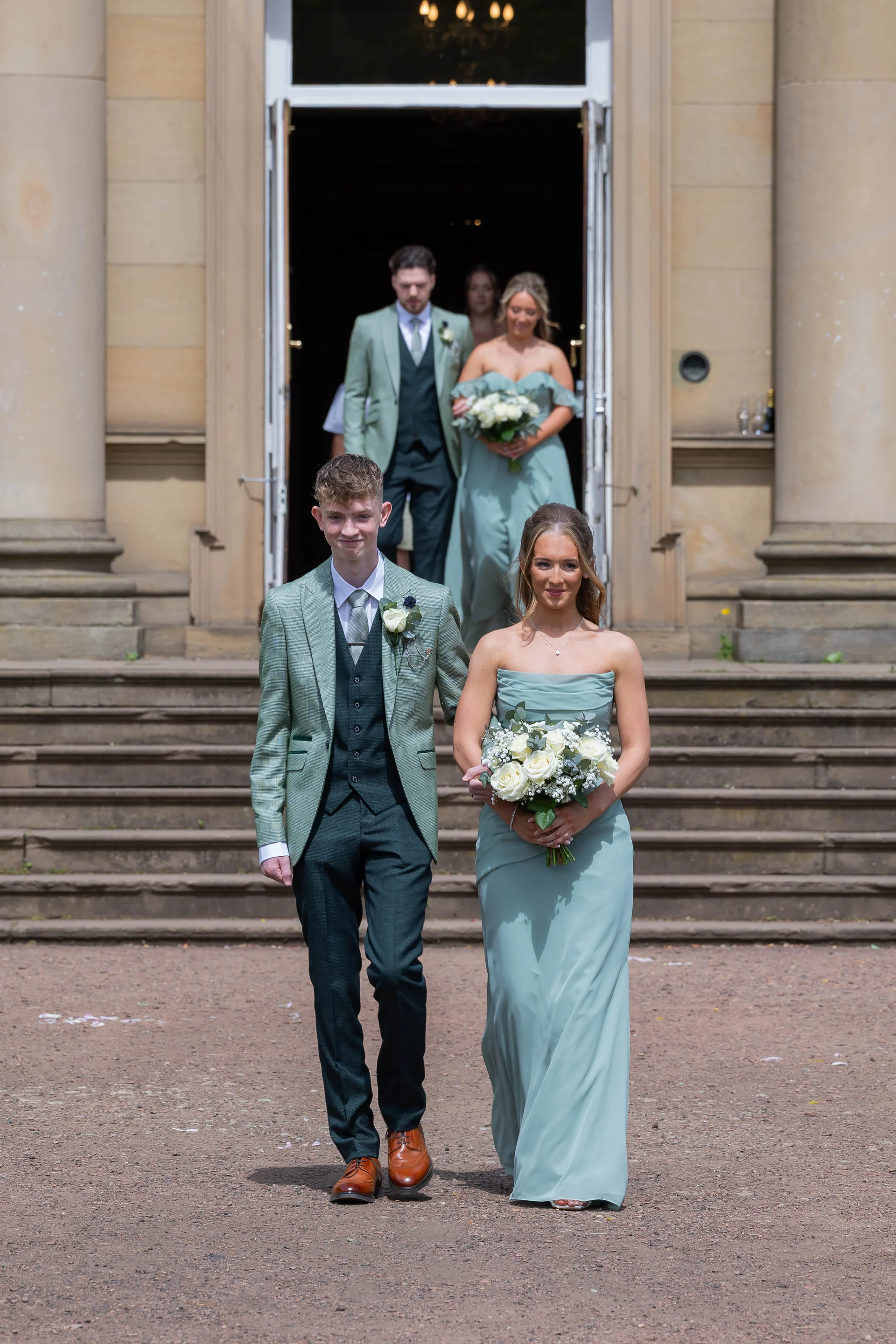 A groomsman in a green suit and waistcoat walks arm-in-arm with a bridesmaid in a sage green strapless dress holding a bouquet of white roses outside a grand country house venue.