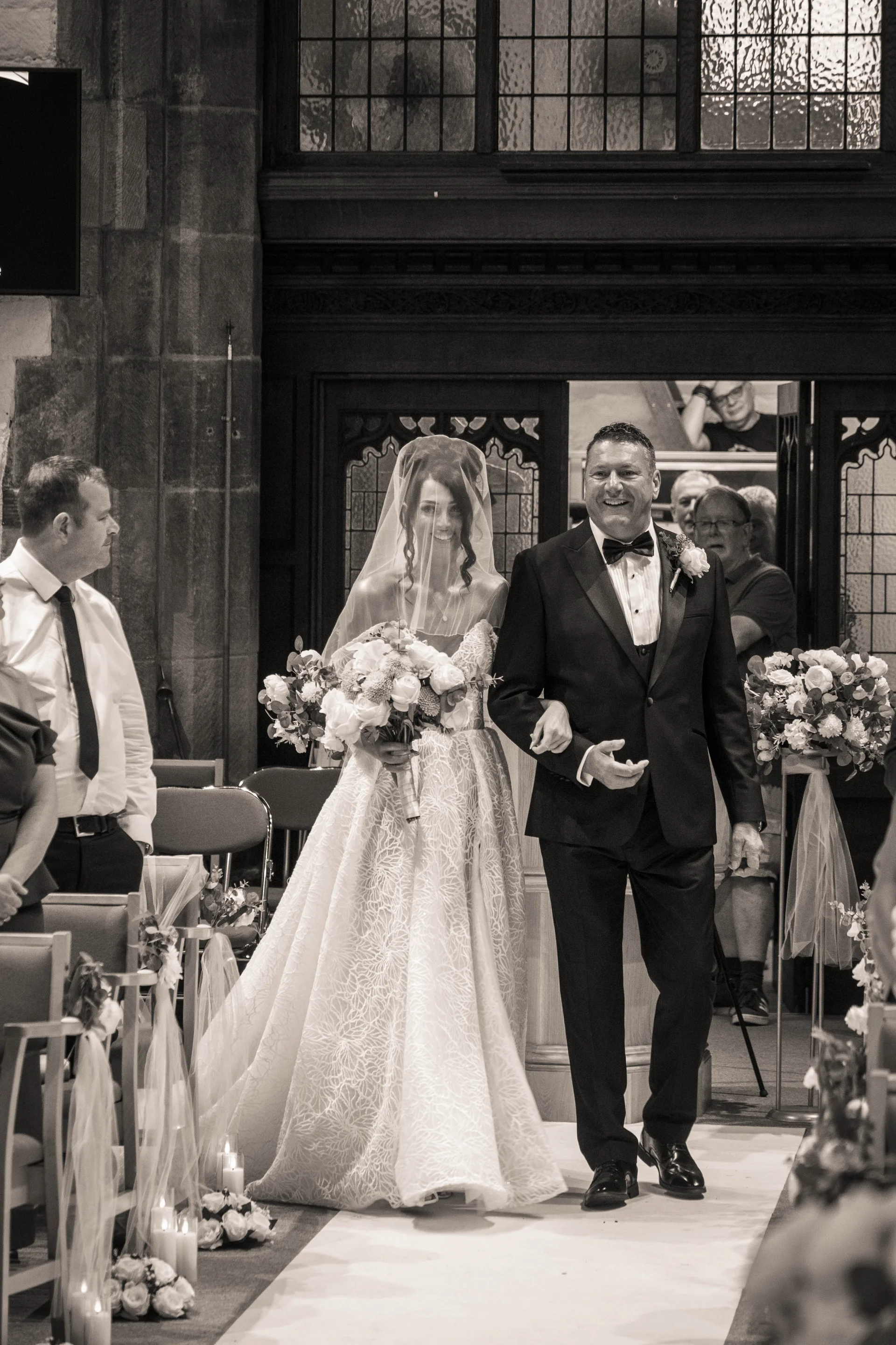 A smiling bride walking down the aisle in a textured ball gown with a blusher veil, linked arm-in-arm with her father in a black tuxedo inside a traditional church.