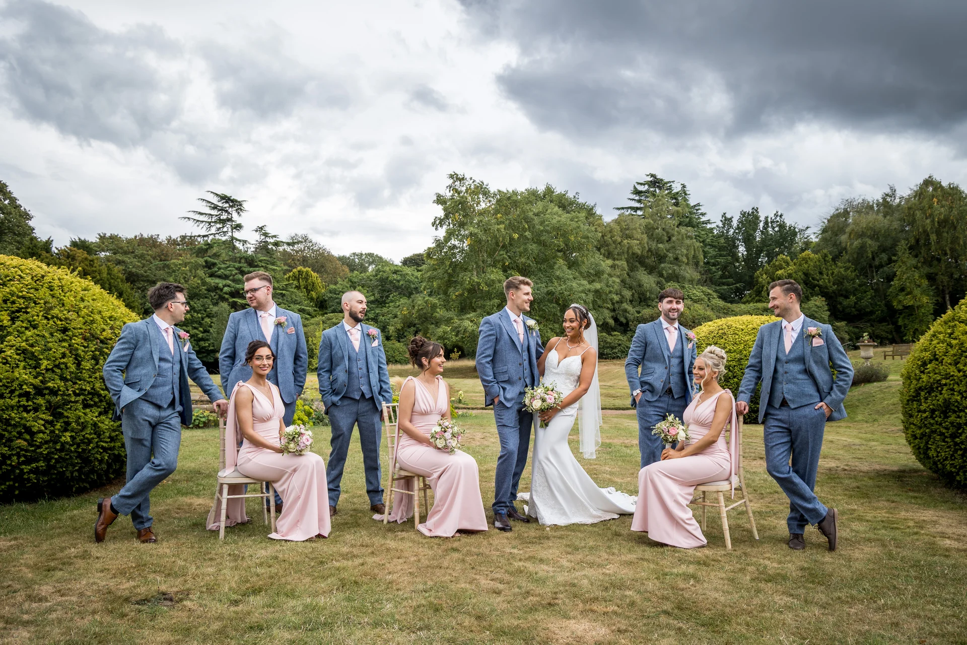 The bride and groom pose happily on a grassy lawn with their wedding party wearing blue suits and pink dresses.