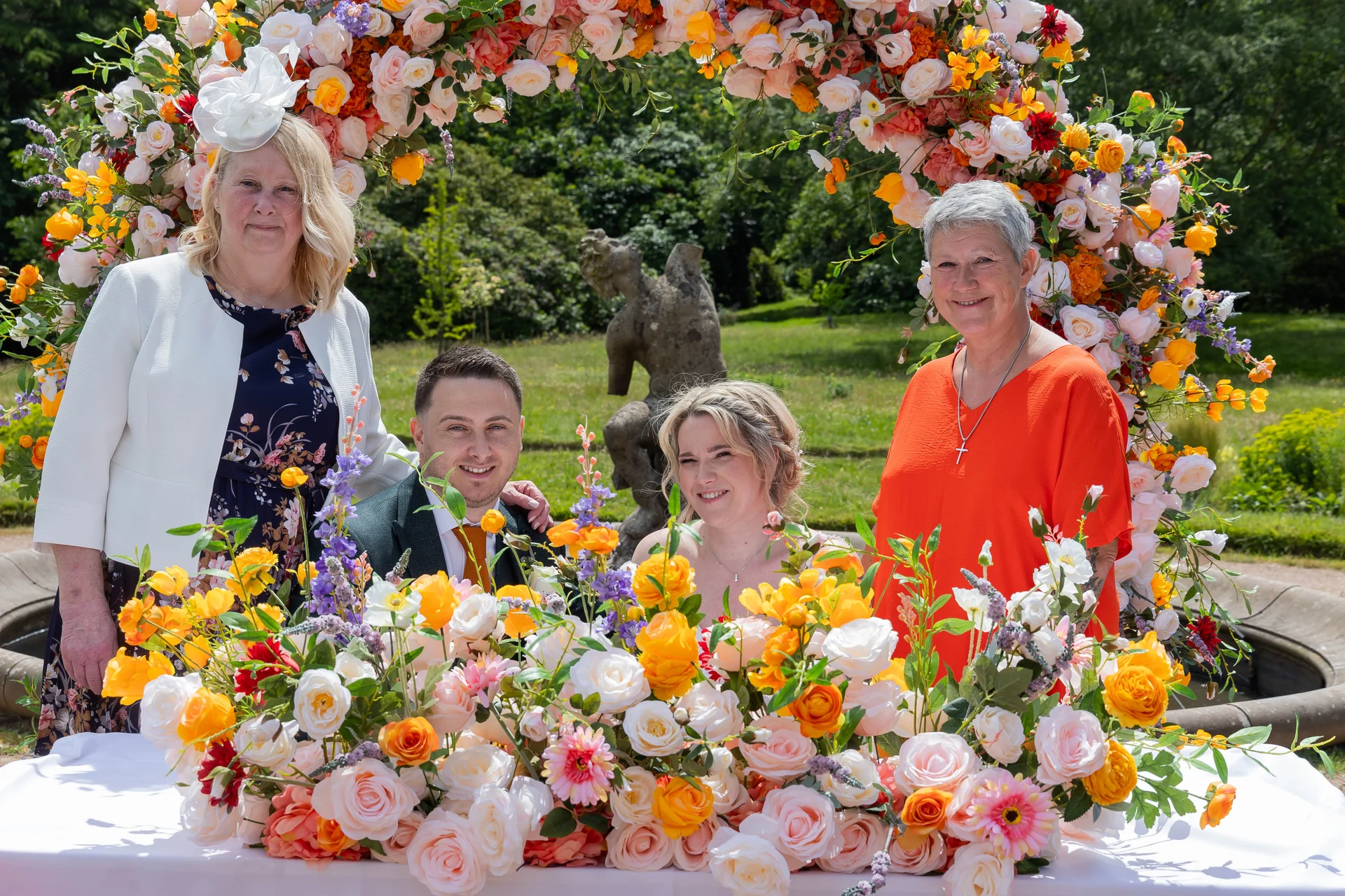 A smiling newlywed couple pose with family members in front of a vibrant, flower-filled circular arch in a sunlit garden setting.