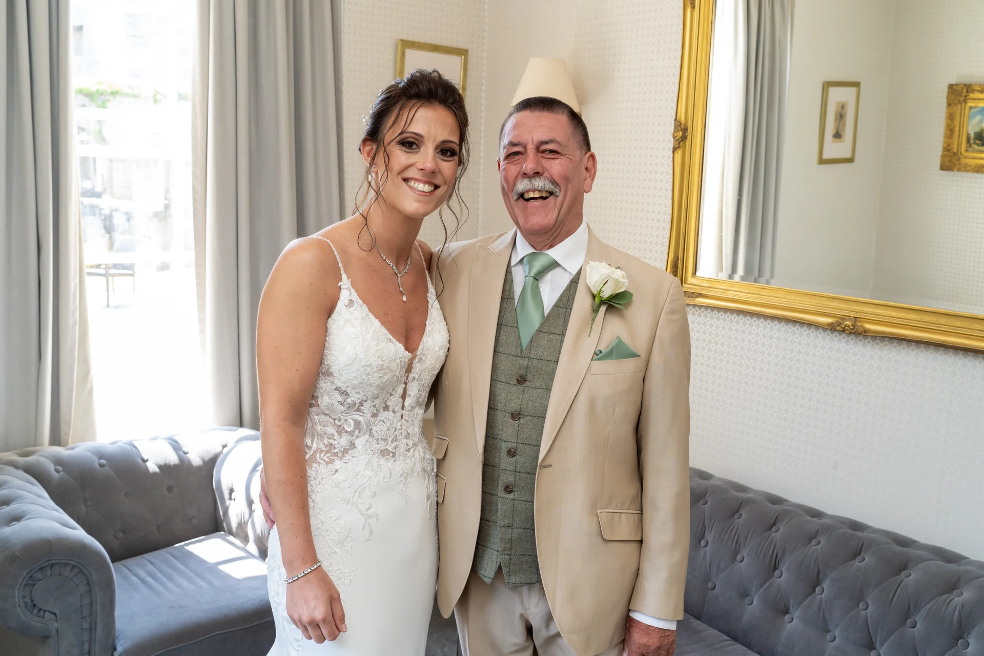 A radiant bride in a lace gown smiles warmly alongside her beaming father in a tan suit and tweed waistcoat.