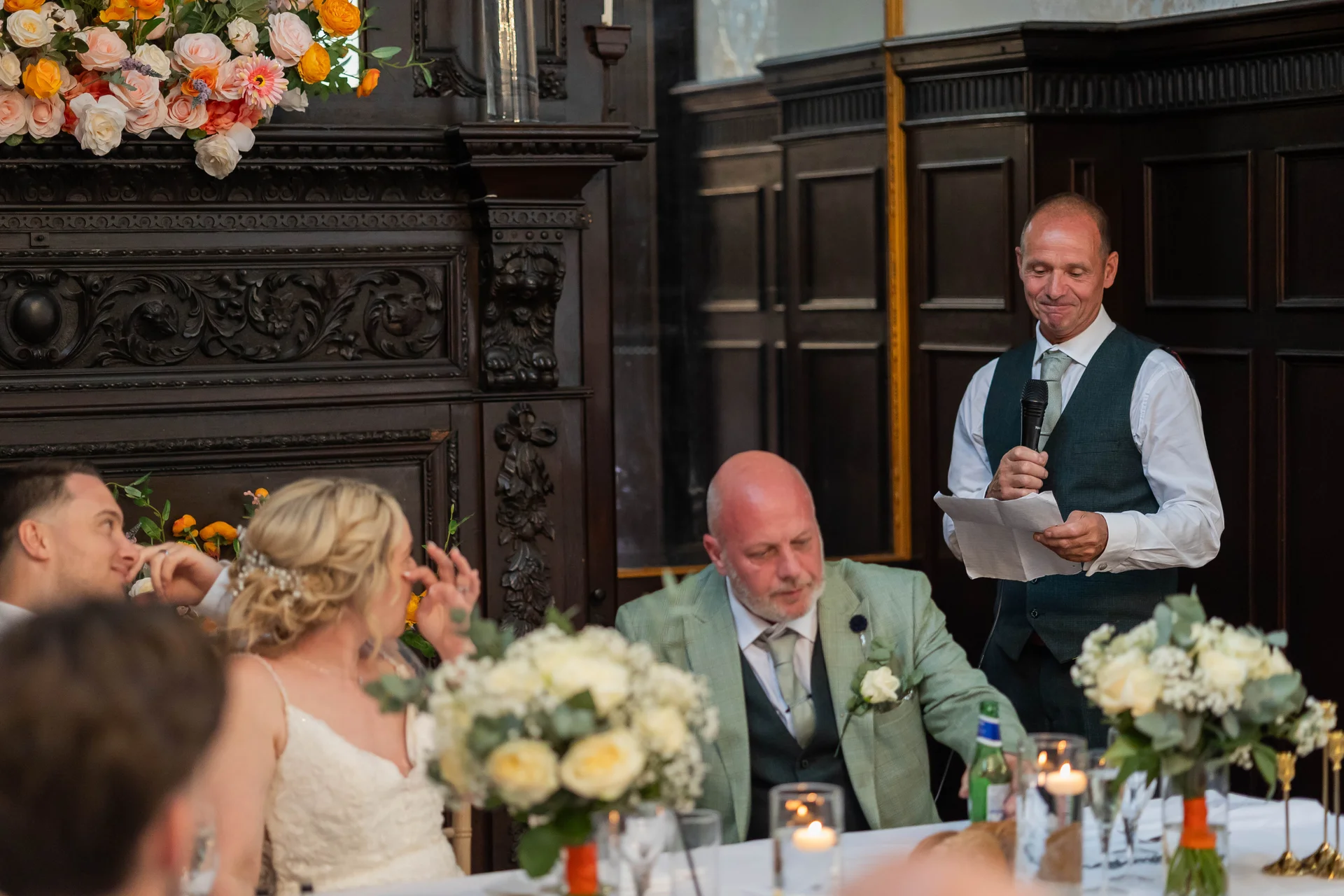 A smiling speaker delivers a wedding speech standing by a grand dark wood fireplace while the bride and guests listen from the top table.