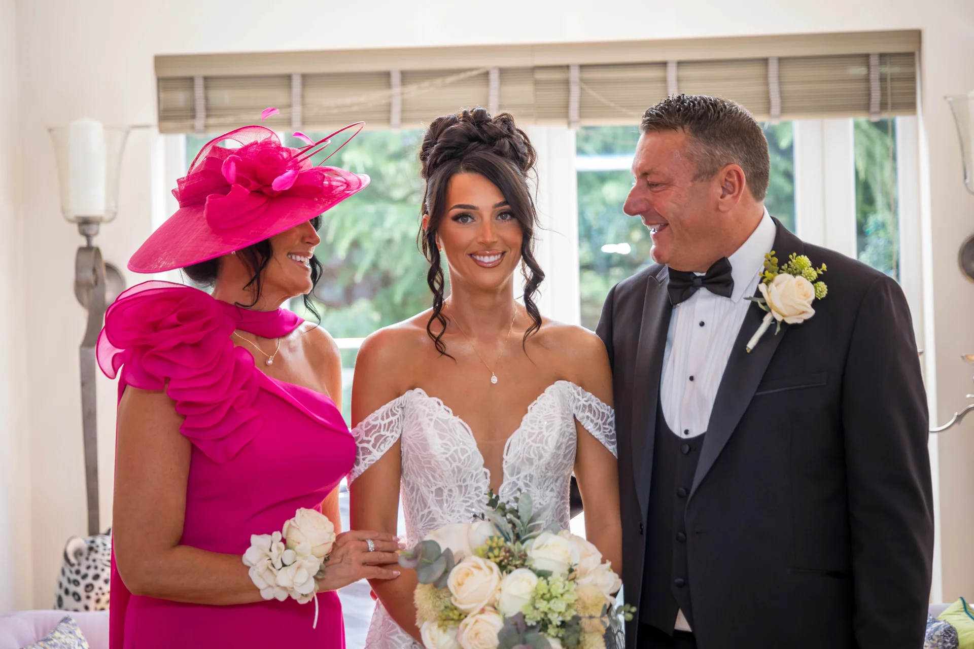 A radiant bride stands smiling between her mother in a bright pink dress and fascinator and her father in a classic black tuxedo during wedding preparations.