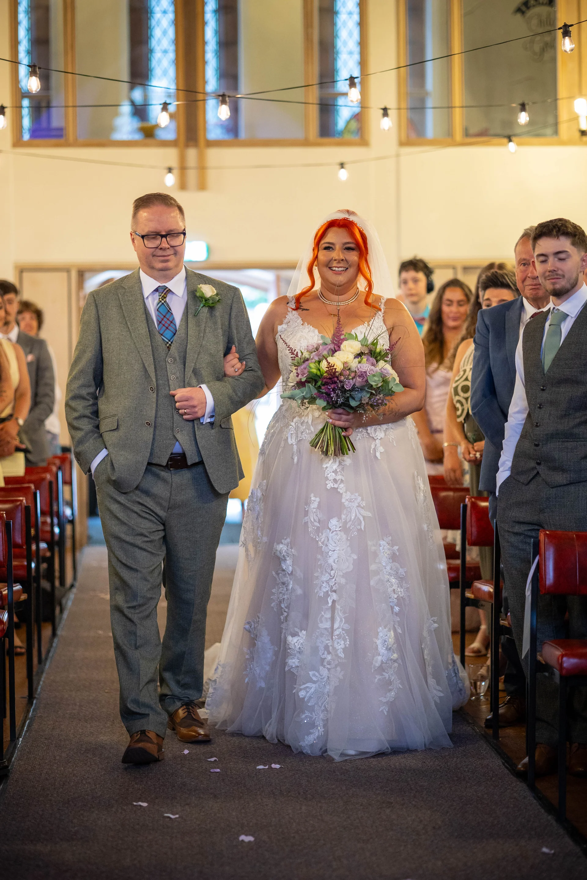 A smiling bride with vibrant red hair walks down the aisle arm in arm with her father, holding a colourful hand-tied floral bouquet in a bright church venue.