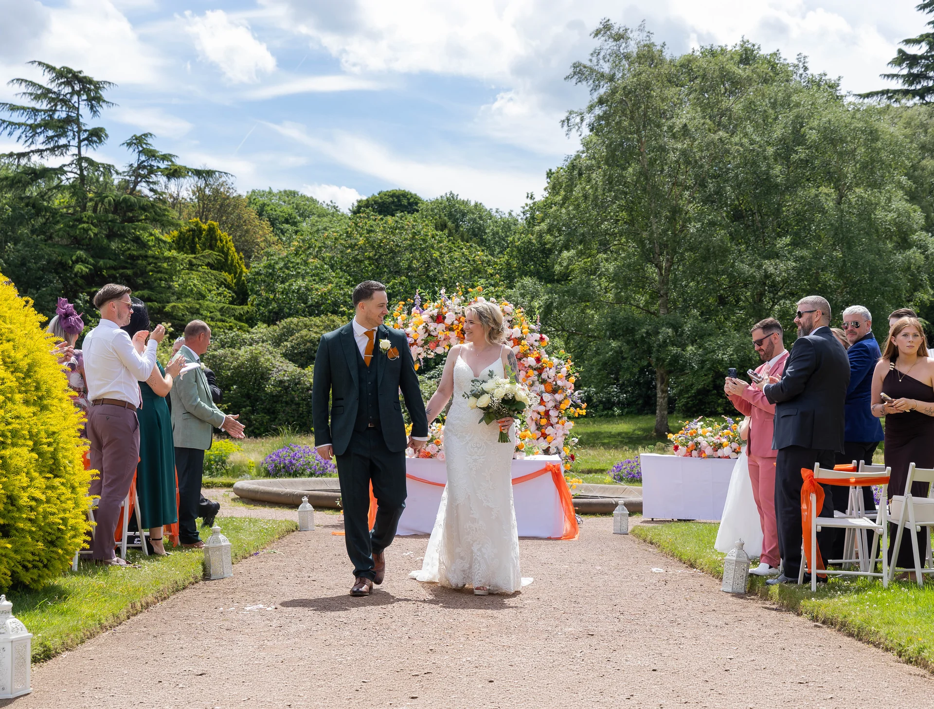 Newlywed bride and groom walk hand in hand down the outdoor gravel aisle, smiling at each other as guests cheer and applaud in a lush garden setting.