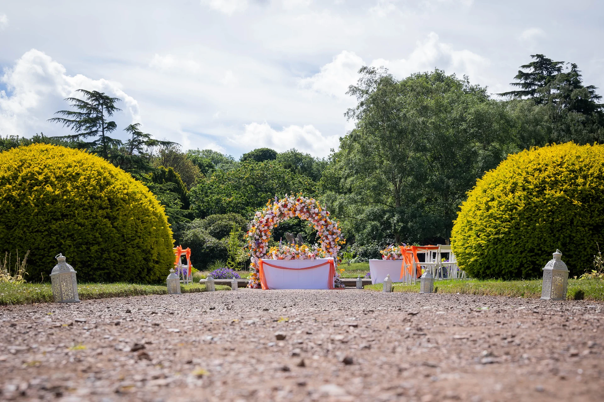 An outdoor wedding ceremony setup featuring a floral circular arch and white lanterns lining a gravel path in a manicured garden under a bright sky.