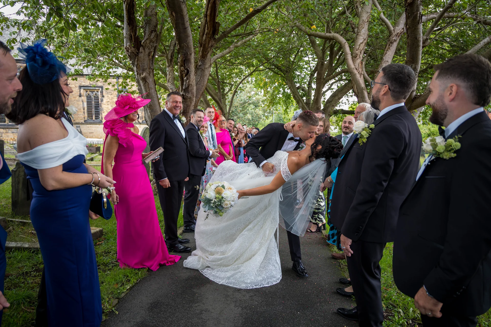 The groom dips his bride for a dramatic kiss on the path outside the church while wedding guests cheer and applaud.