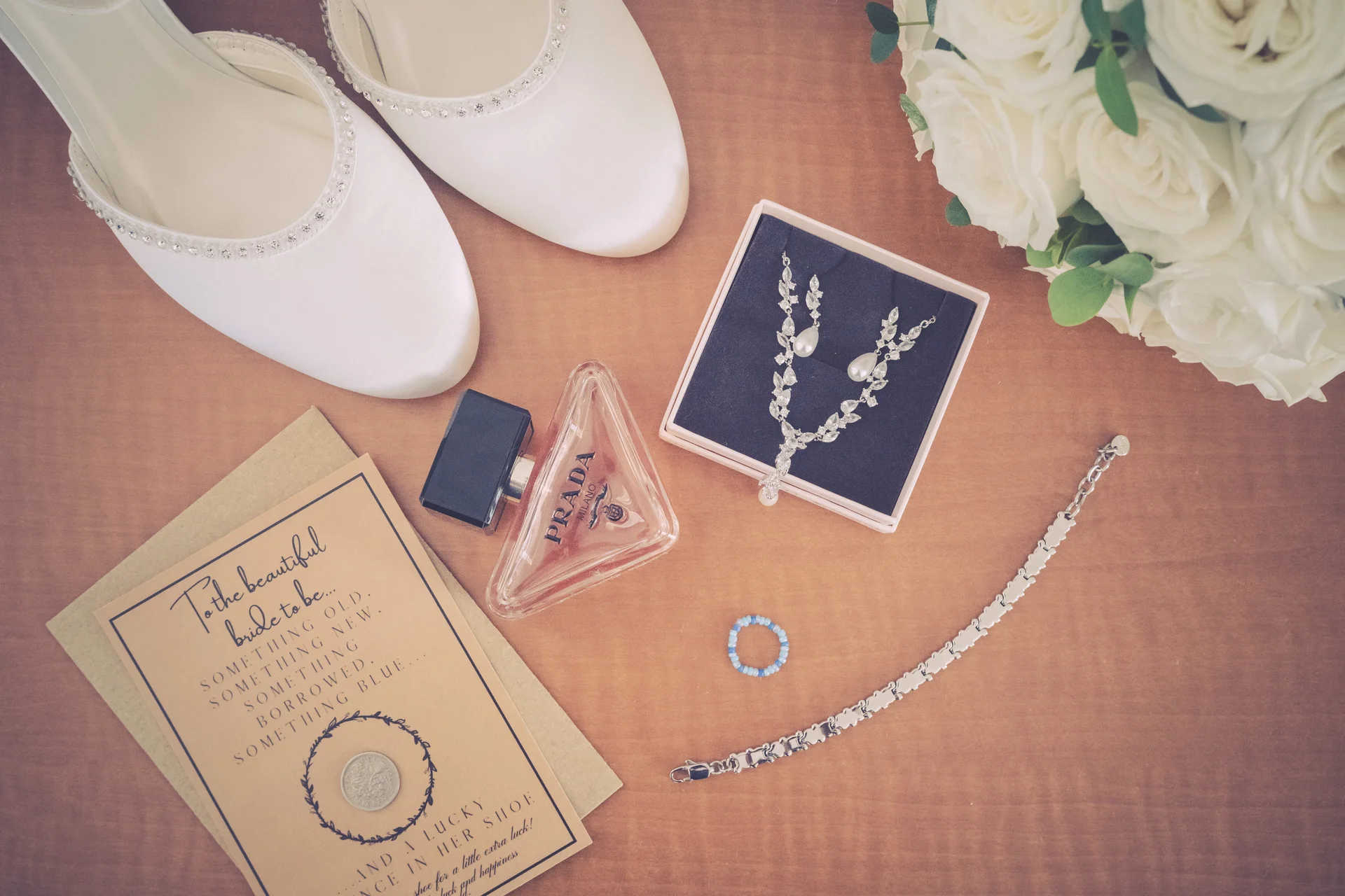 An elegant bridal flat lay featuring white satin shoes, a Prada perfume bottle, delicate jewellery in a presentation box, a handwritten card with a lucky sixpence, and a bouquet of white roses.