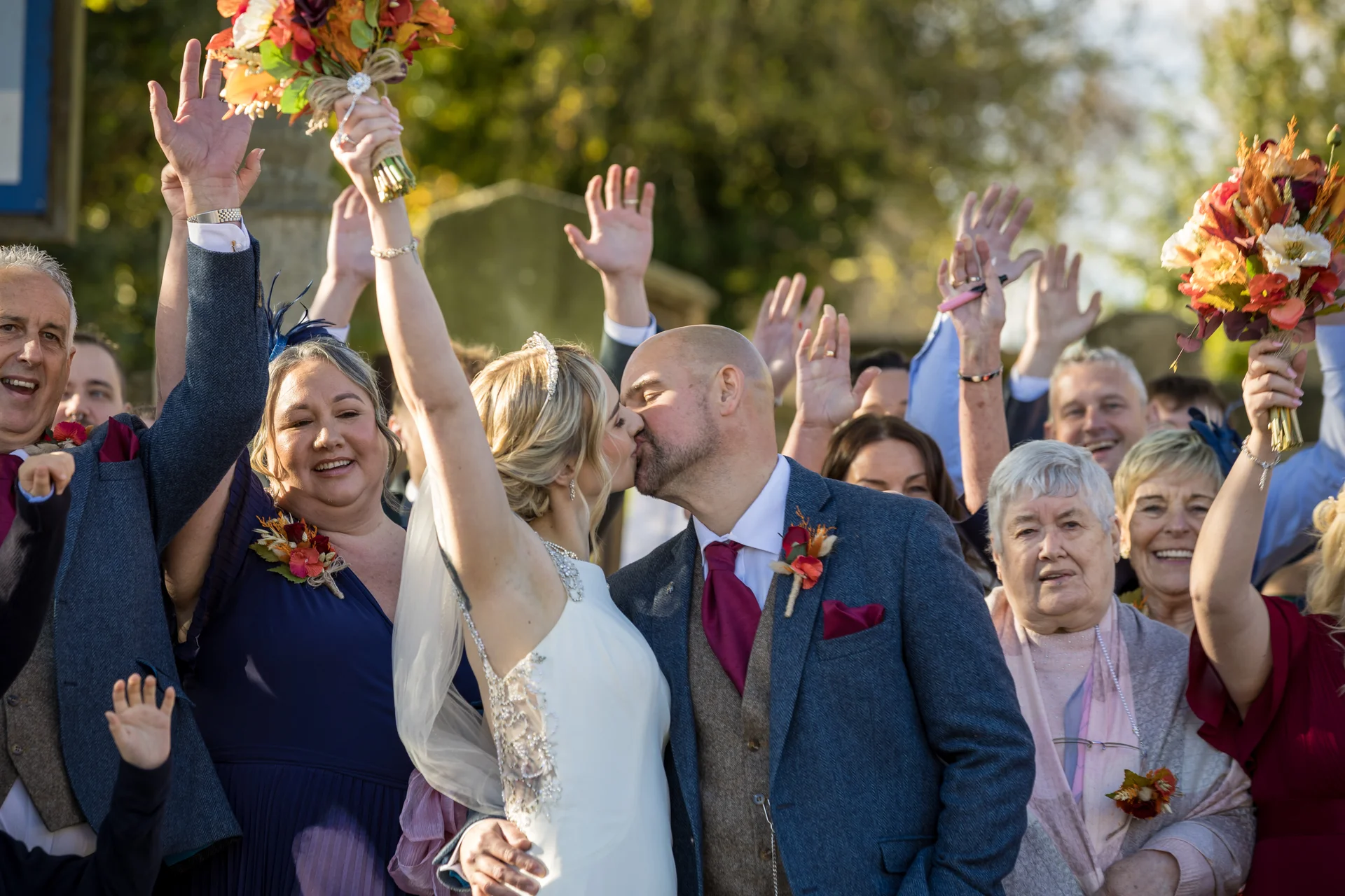 The bride and groom share a passionate kiss as family and friends raise their hands and bouquets in celebration outdoors during an autumn wedding.
