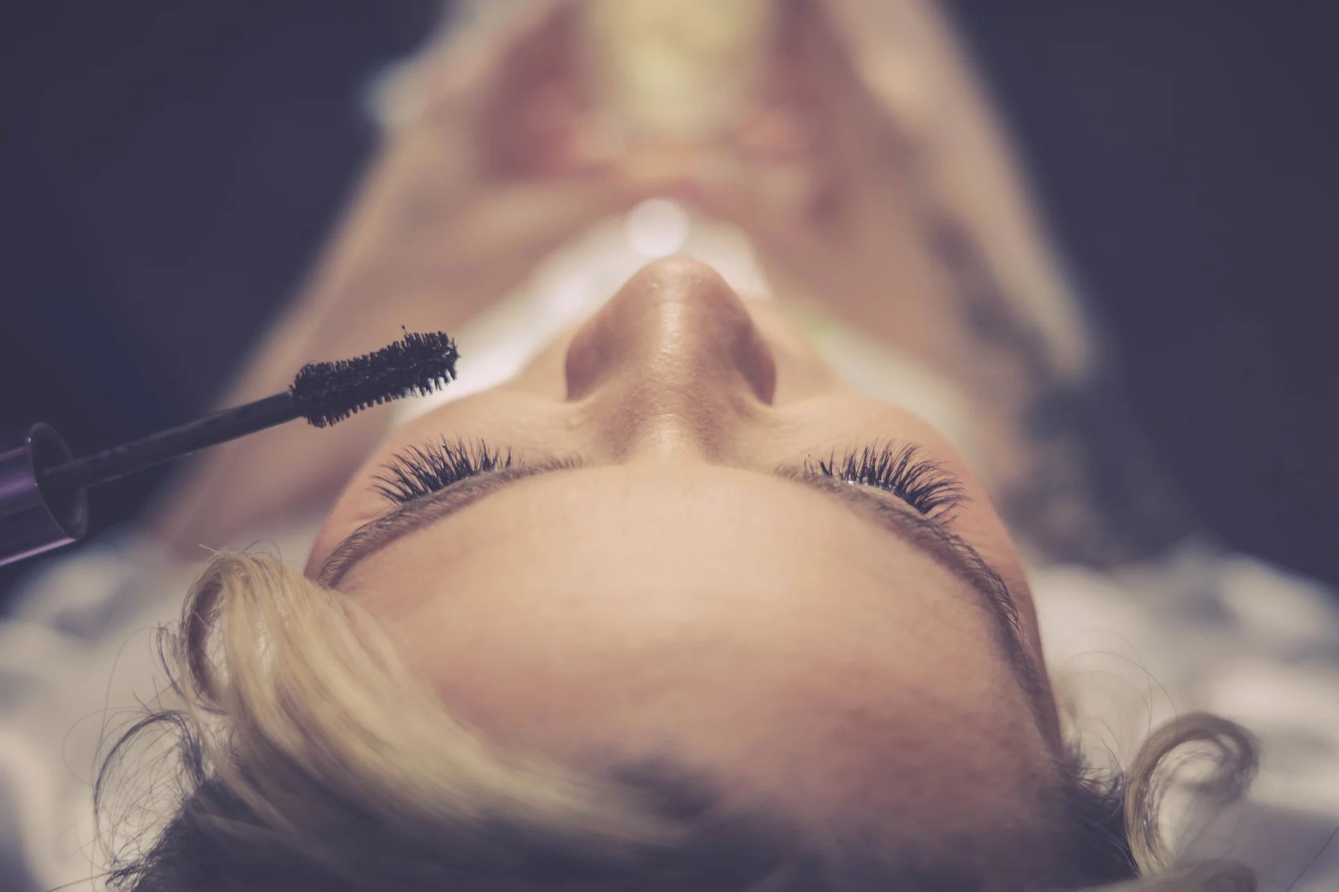 A close-up shot from above captures a bride having mascara applied to her eyelashes during morning wedding preparations.