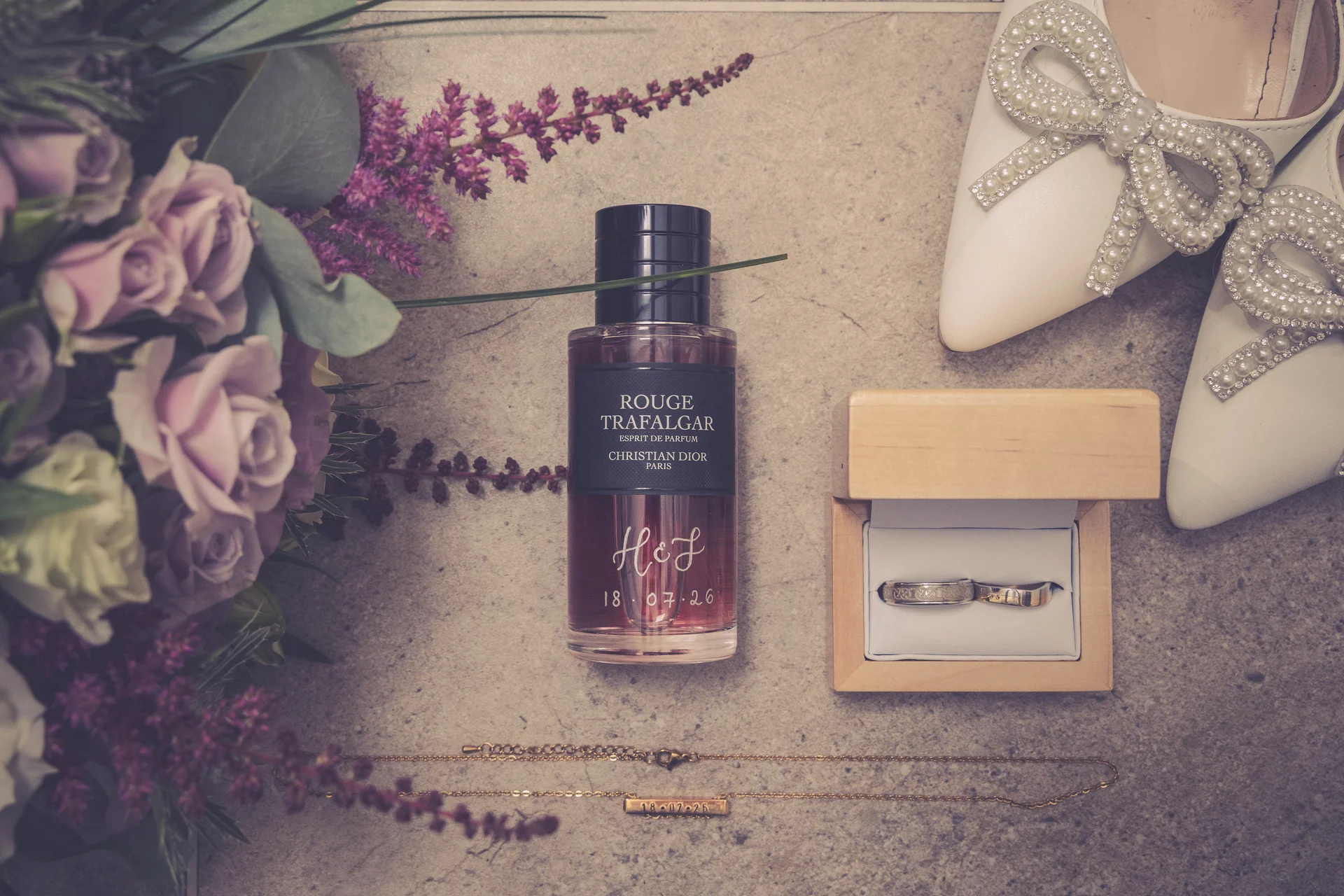 A bridal flat lay featuring a perfume bottle, wedding bands in a wooden box, pearl-embellished bow heels, a gold necklace, and a floral bouquet with purple roses.