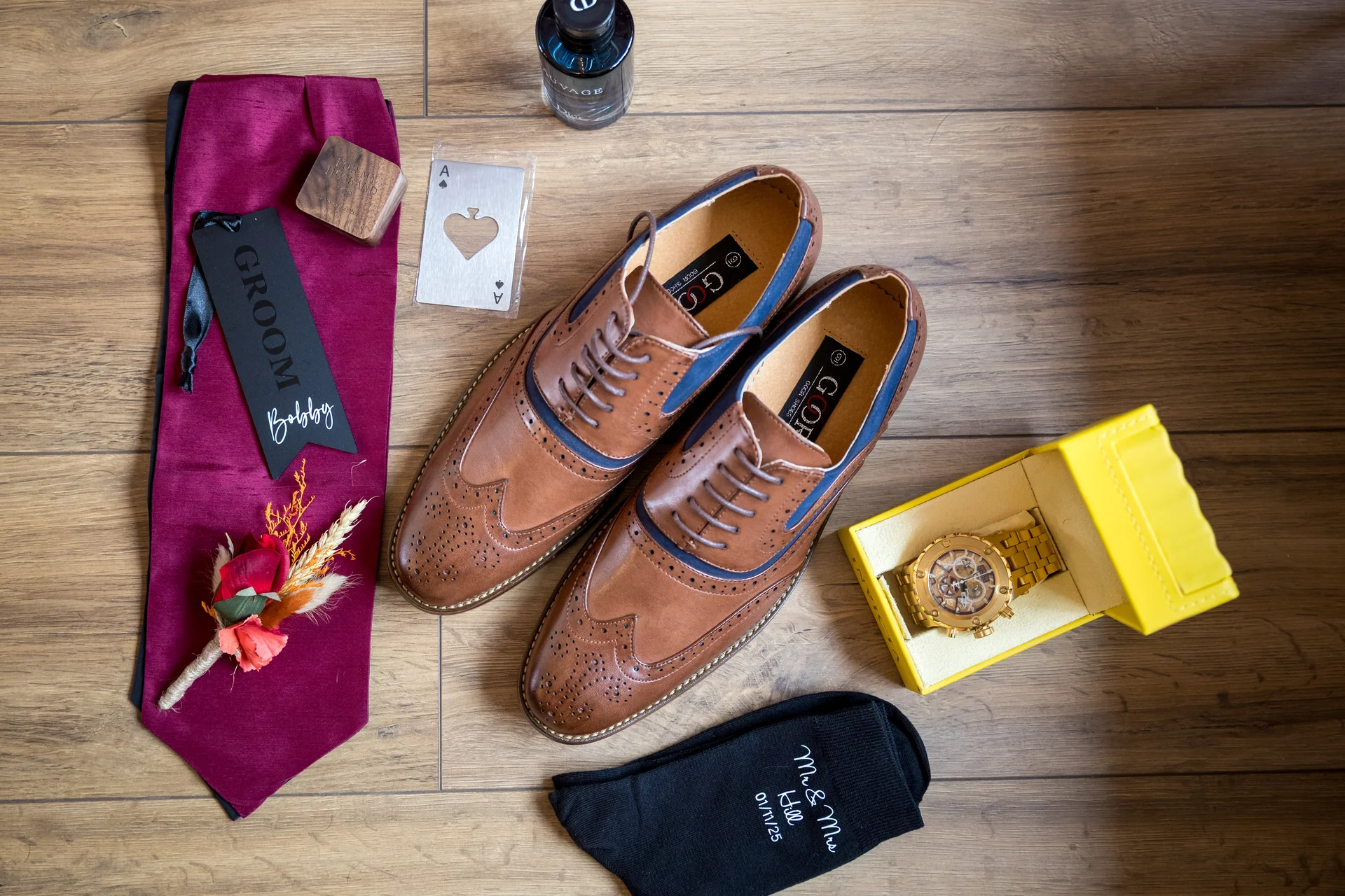 An overhead flat lay captures a groom's wedding morning accessories, including brown brogues, a burgundy tie, a floral buttonhole, a gold watch in its yellow box, and a pair of personalised black socks on a wooden floor.