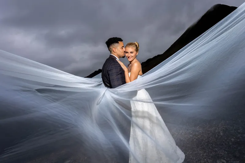 A newlywed couple embraces outdoors against a dramatic mountain landscape under stormy grey skies, framed by a massive flowing veil catching the wind.