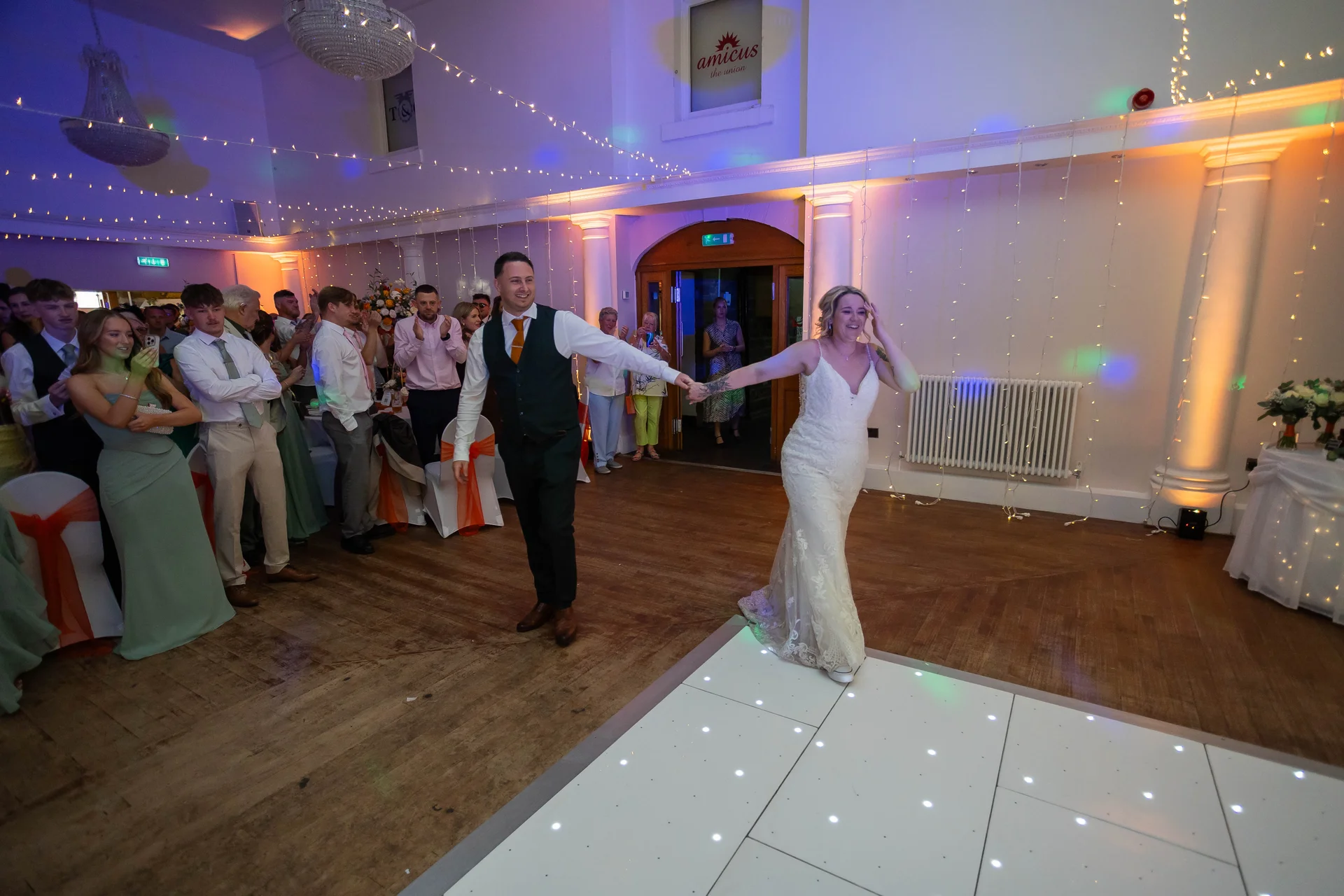 The bride and groom joyfully walk onto the illuminated white dance floor hand in hand during their evening wedding reception as guests applaud in the background.