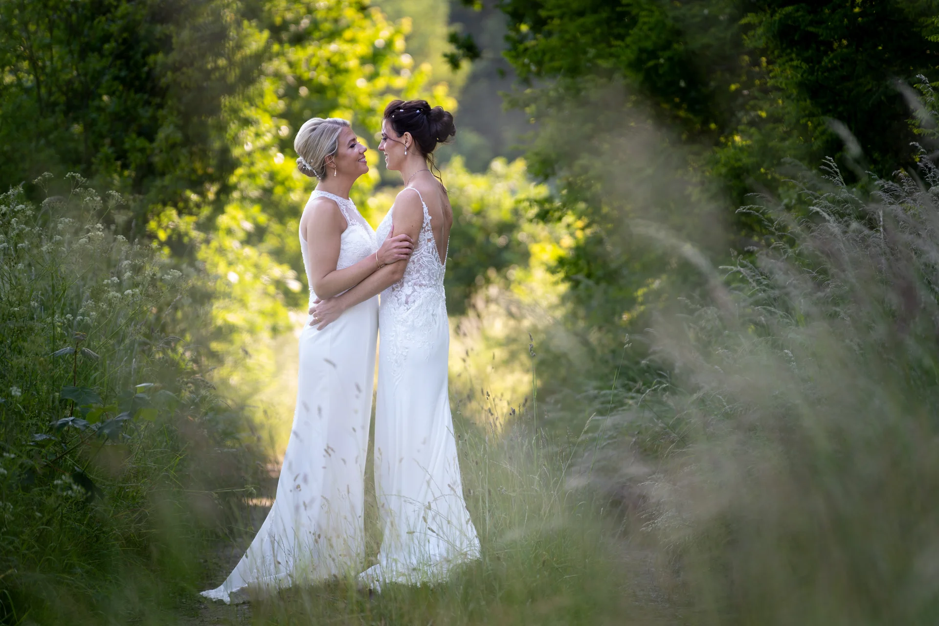 Two brides embrace tenderly on a sunlit woodland path framed by tall grass and lush green foliage during golden hour.
