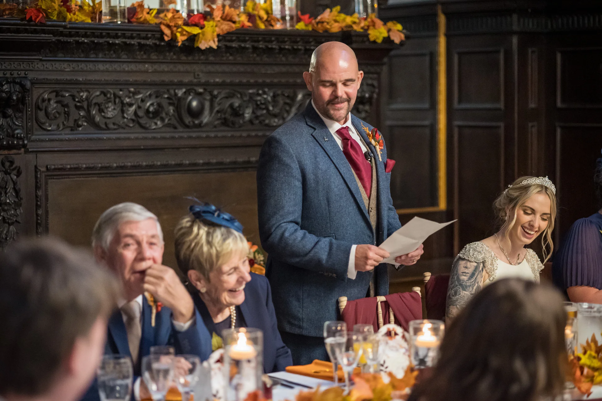 A groom stands delivering his speech during the wedding breakfast, holding notes while seated guests laugh and enjoy the moment in an autumnal candlelit venue.