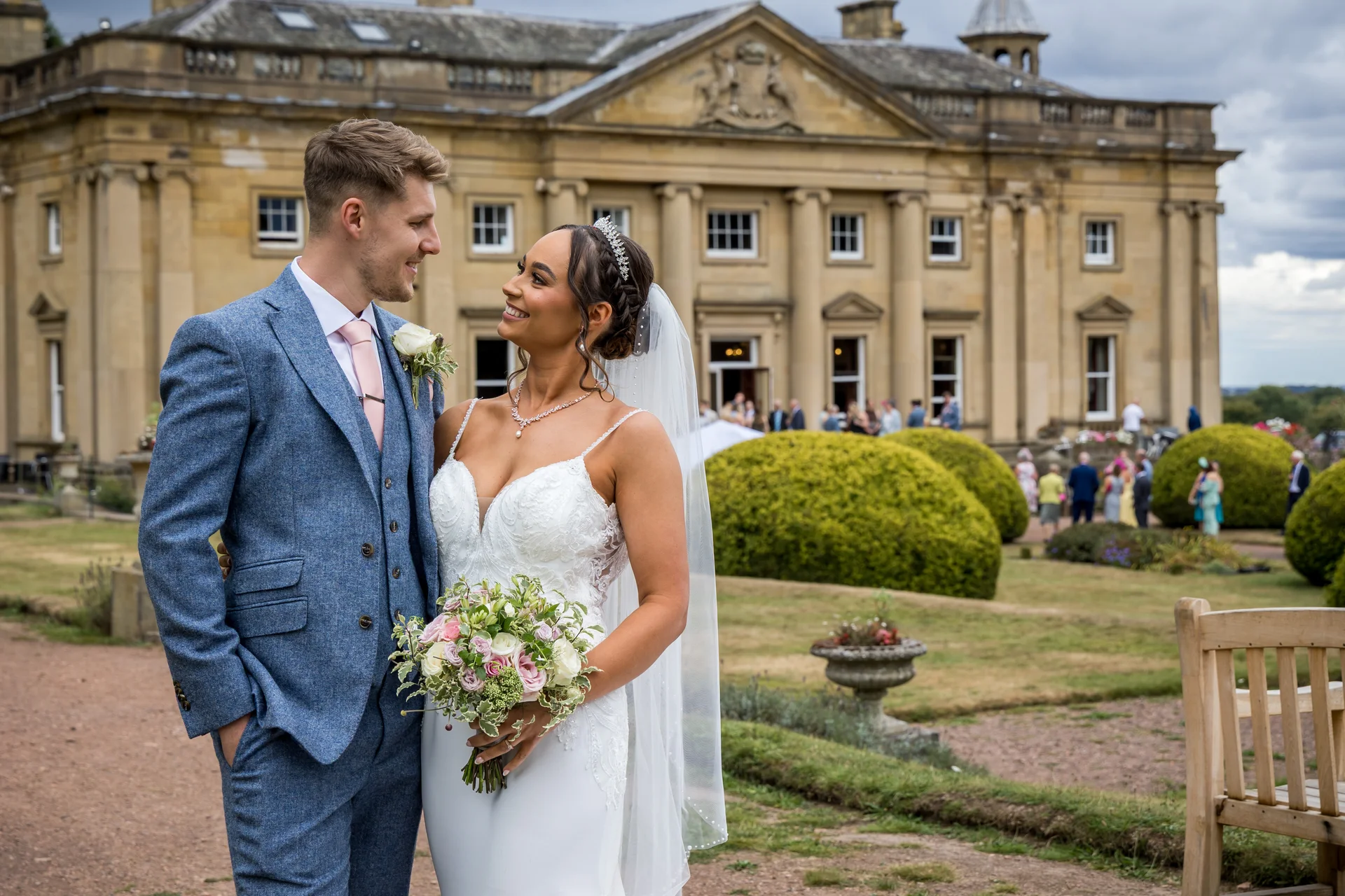 A smiling bride and groom pose for a portrait outside a grand country house, with the bride holding a mixed floral bouquet and wearing a white wedding dress with a veil.