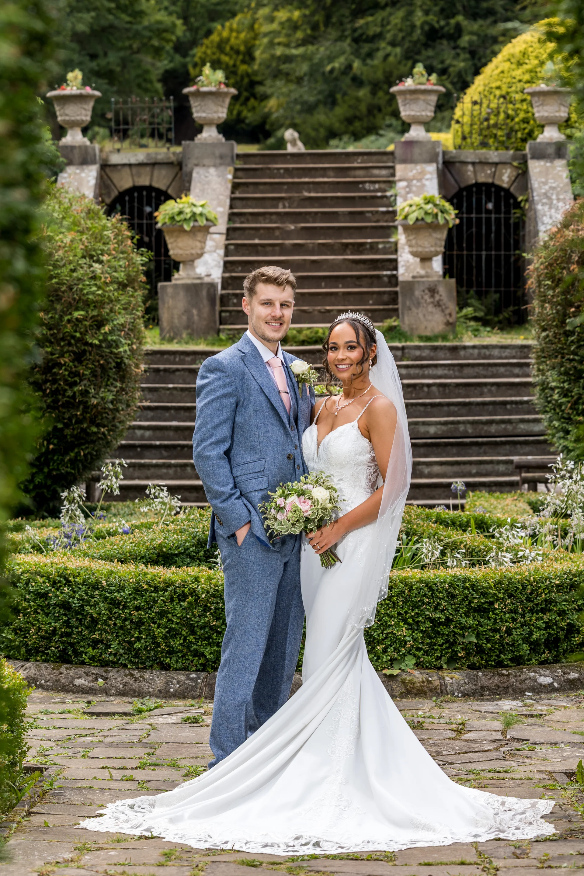 A smiling bride and groom pose in a lush manicured garden with a grand stone staircase and urns in the background.