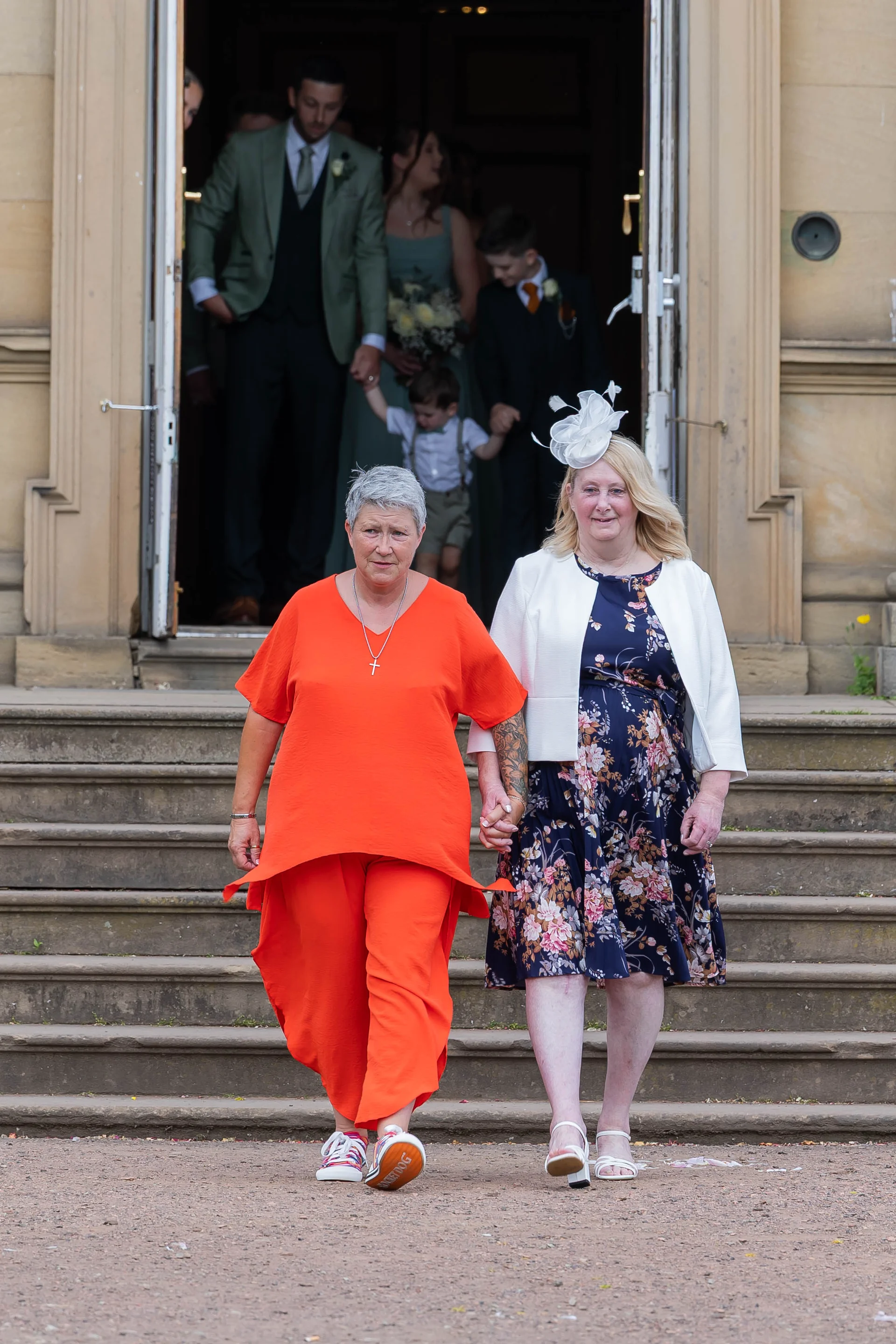 An older same-sex couple walks hand in hand down stone steps outside a country house venue, accompanied by wedding guests emerging from the doorway behind them.