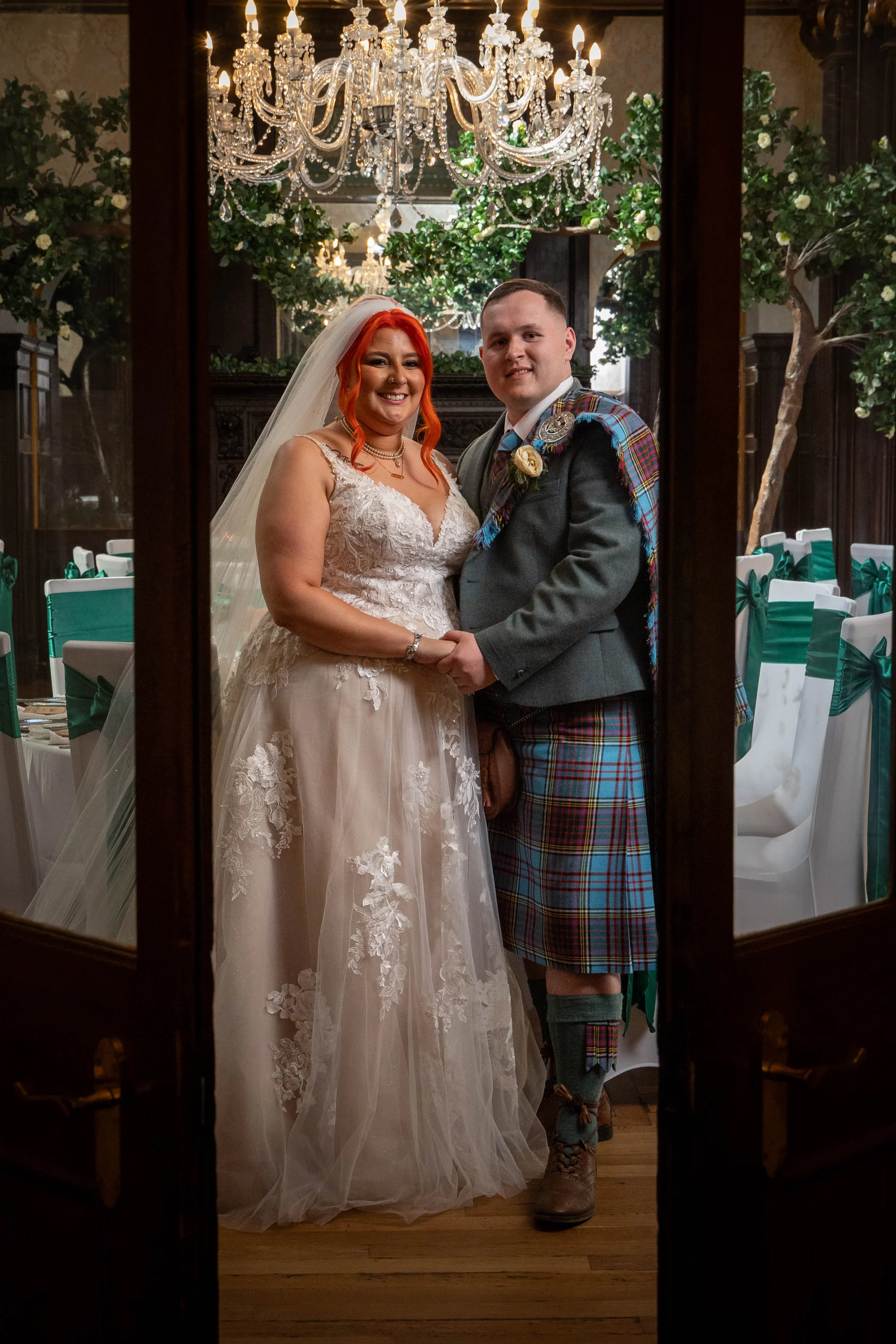 A smiling bride in a lace gown and veil holds hands with her groom dressed in a traditional Scottish kilt inside an elegant venue illuminated by a grand crystal chandelier.