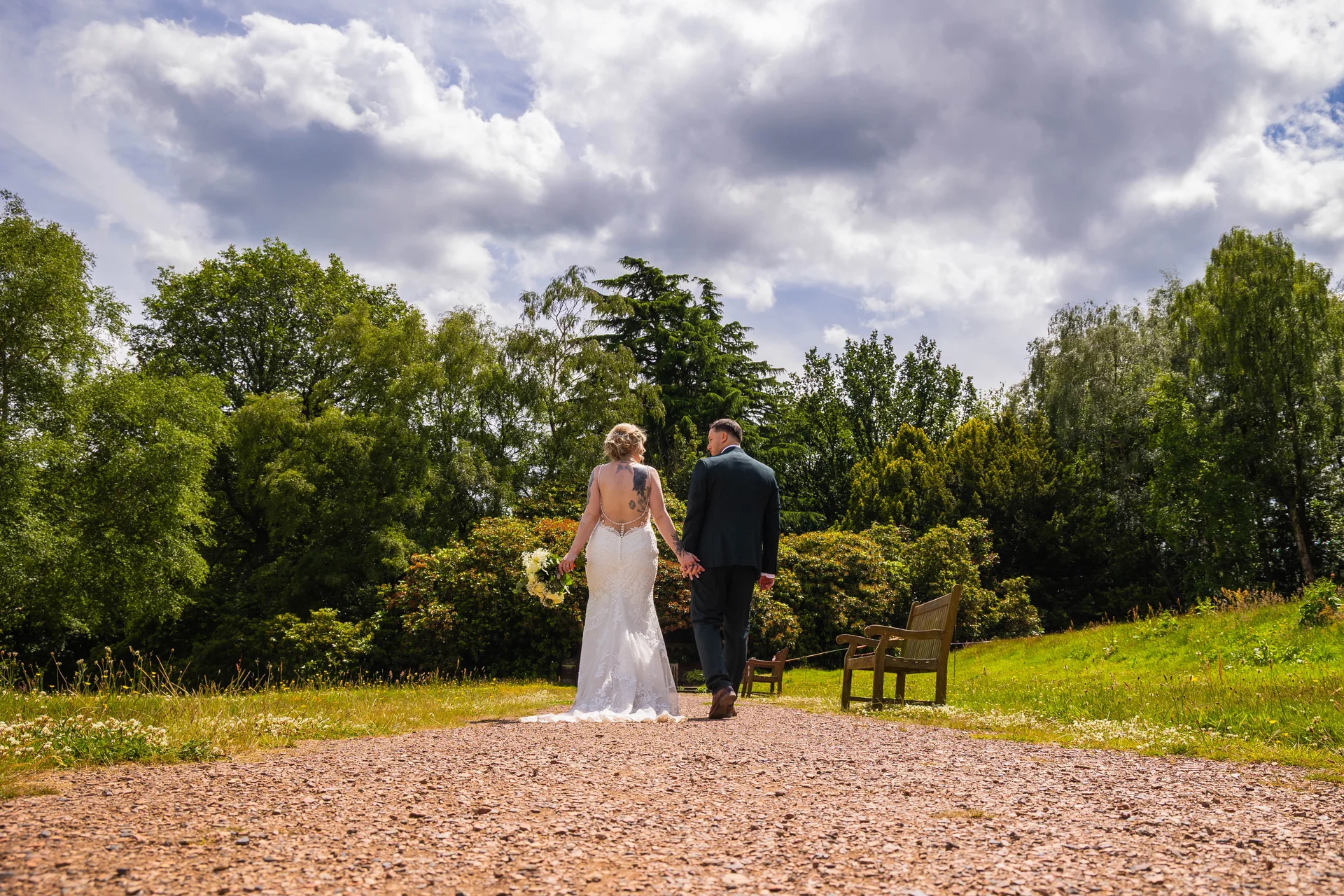 A newlywed couple walks hand in hand along a gravel path in a lush garden setting beneath a dramatic cloudy sky.