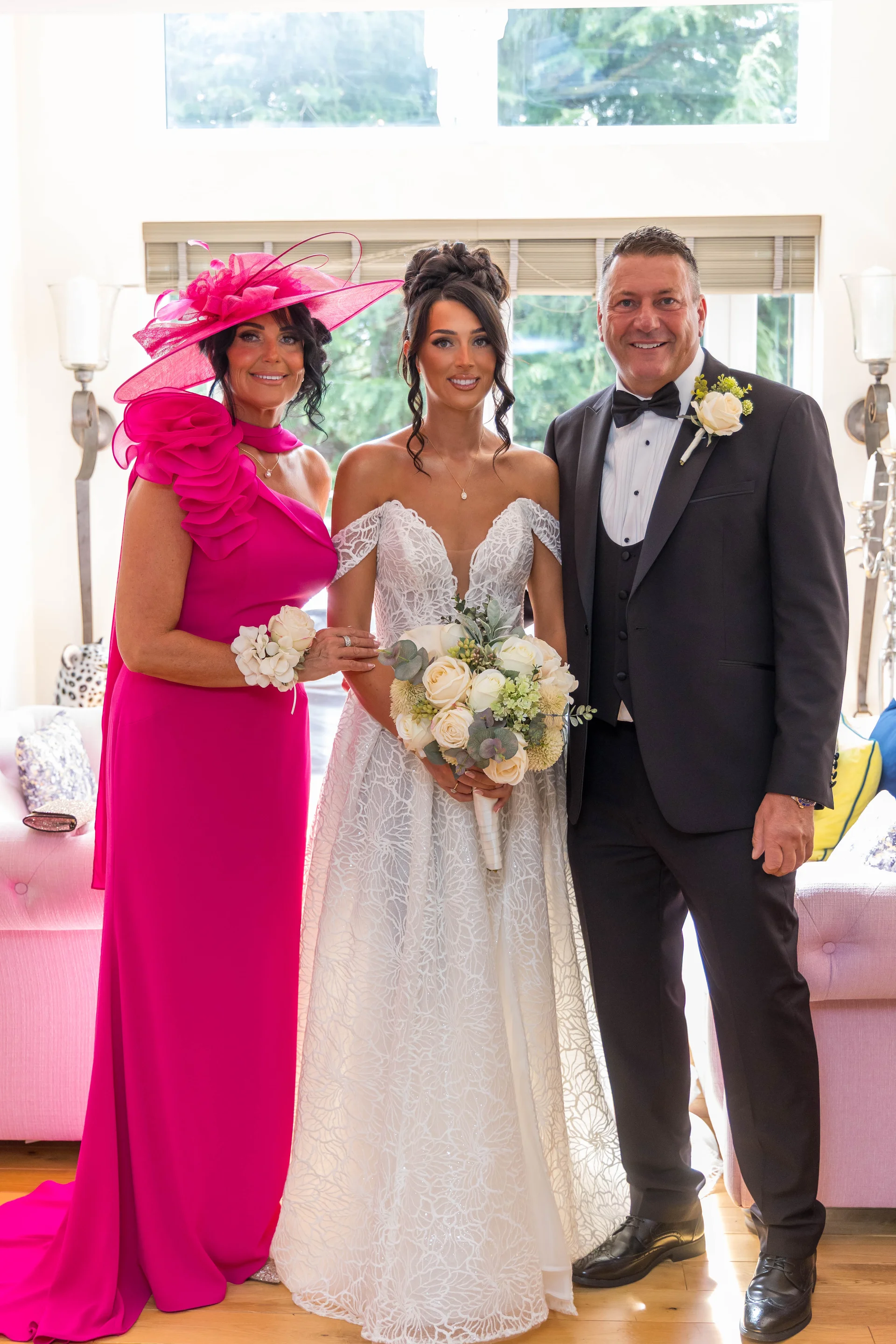 A smiling bride stands proudly between her mother, who wears a vibrant pink gown and matching fascinator, and the groom in a classic black tuxedo.