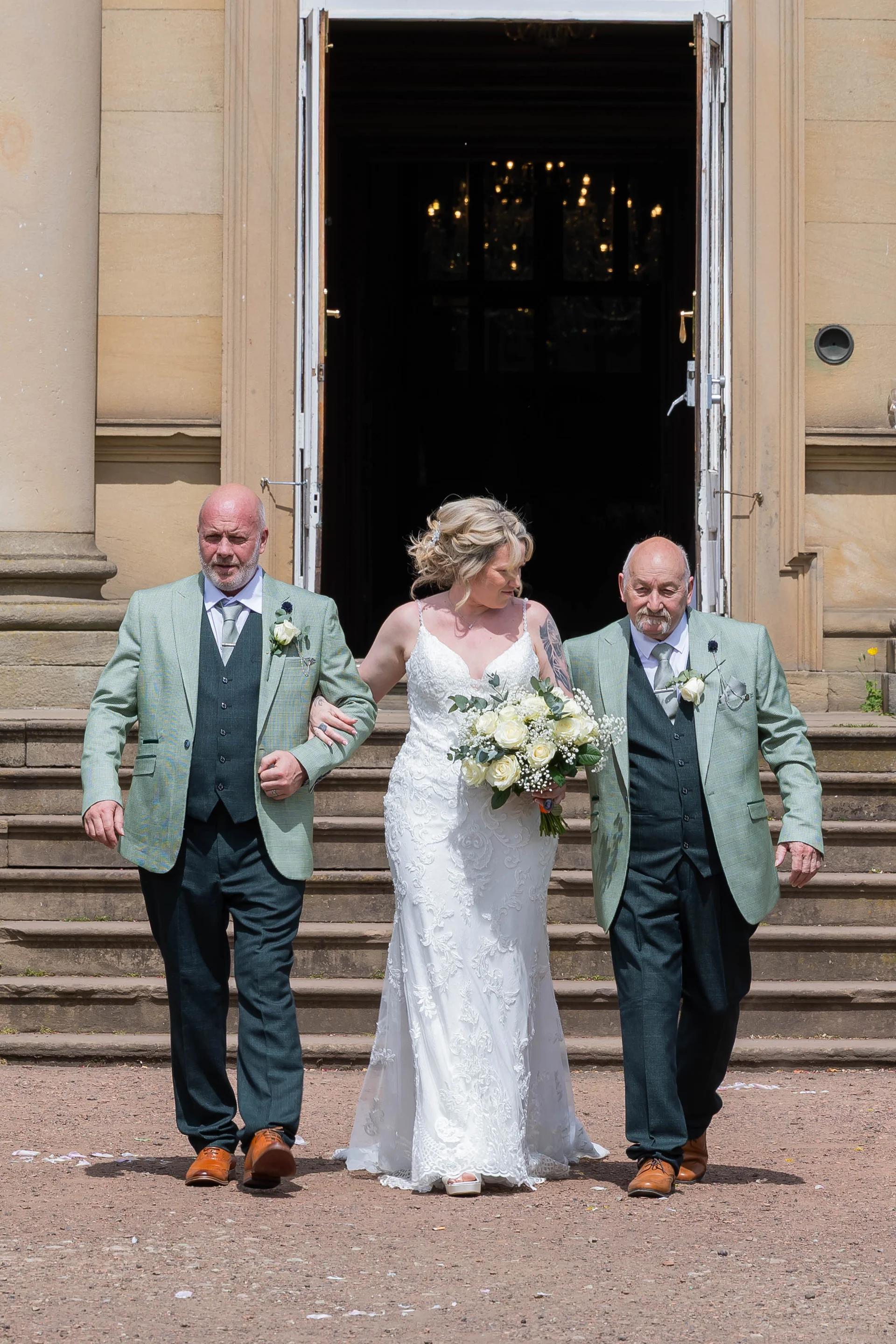 A smiling bride walking arm in arm down stone steps with two family members outside a grand venue, holding a white rose bouquet.