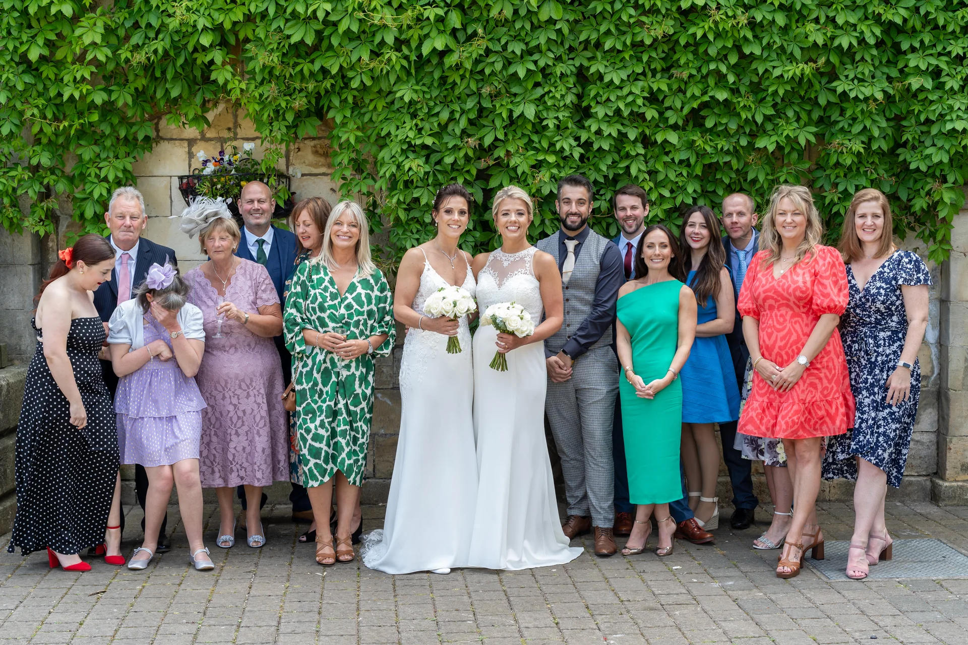 A large wedding party and family group pose smiling and laughing outdoors in front of a stone wall covered in lush green ivy.