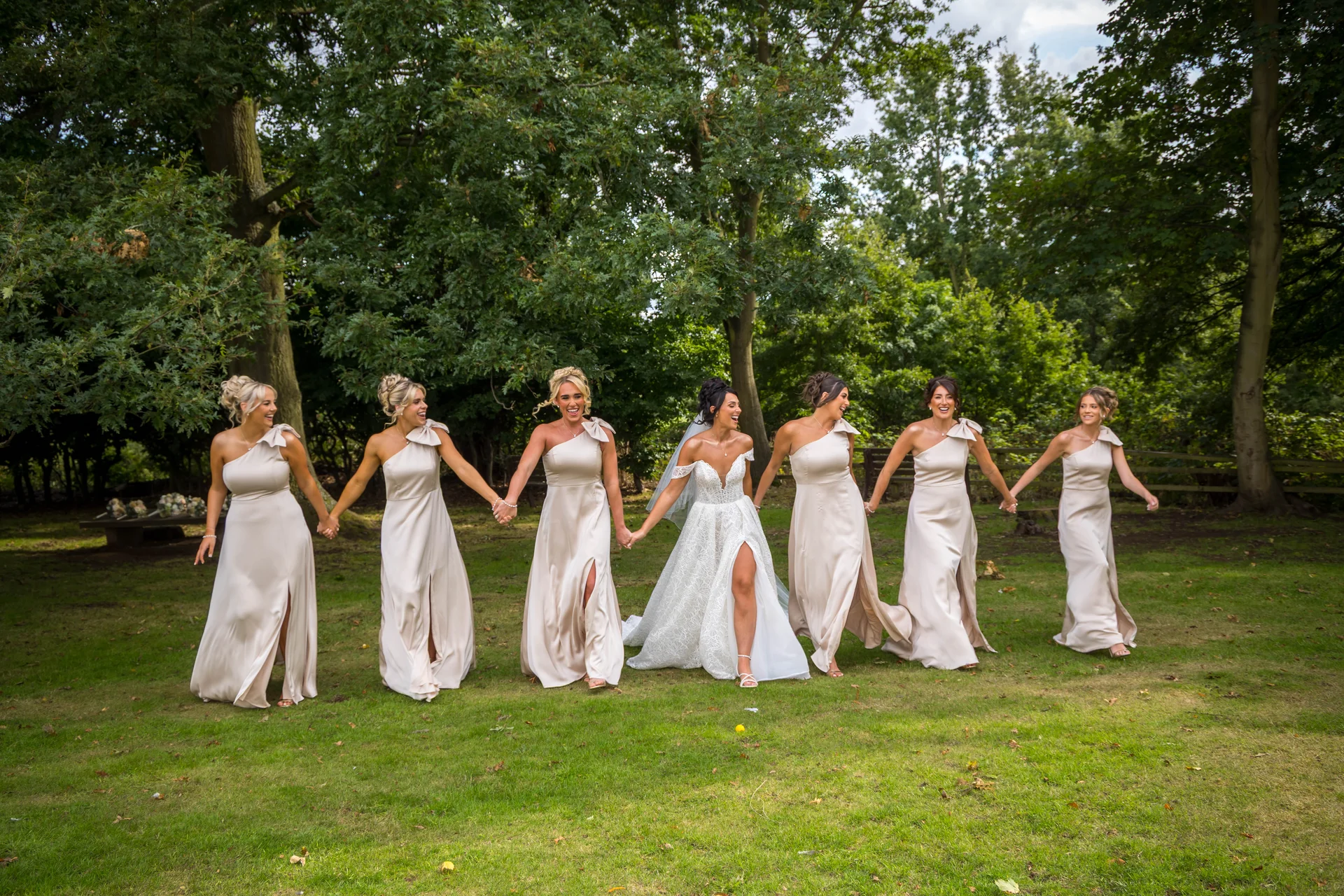 A joyful bride in a white lace gown with a thigh split walks hand in hand through a grassy woodland with six bridesmaids wearing matching one-shoulder champagne satin dresses.