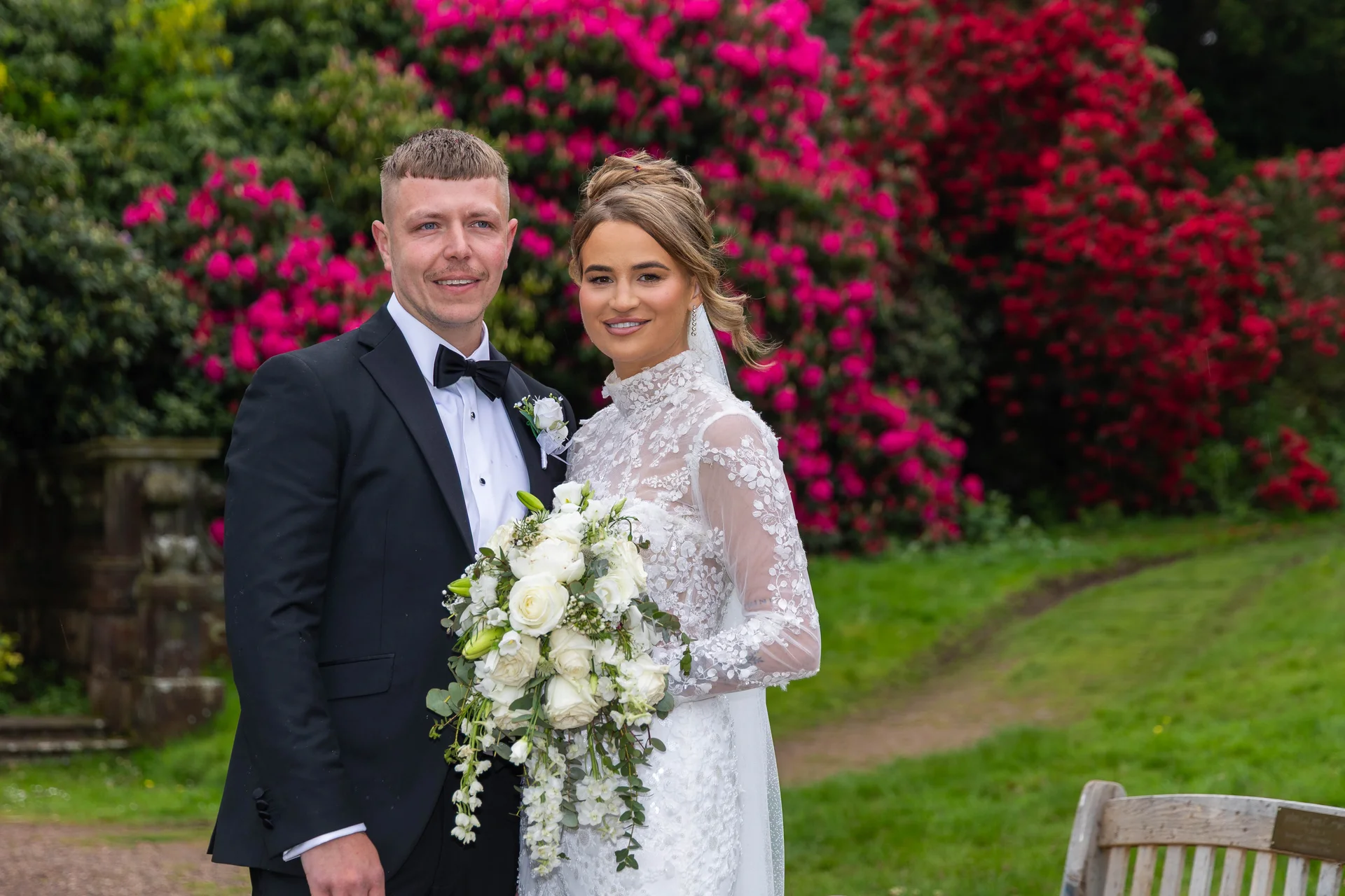 A newlywed couple poses together outdoors in a lush garden, with the bride holding a cascading white floral bouquet and wearing a high-necked lace gown, while the groom wears a classic black tuxedo with a bow tie.