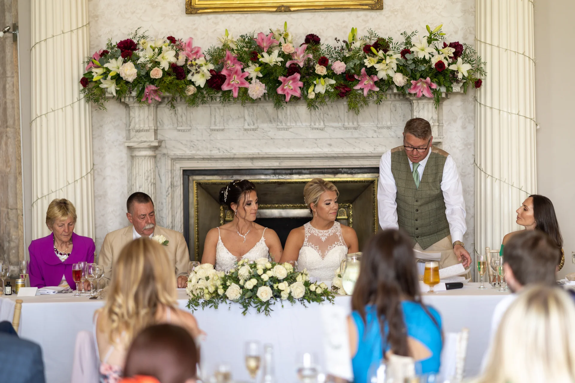 A man stands delivering a speech at the top table during a wedding reception, flanked by two brides and seated guests in front of a grand decorated fireplace.
