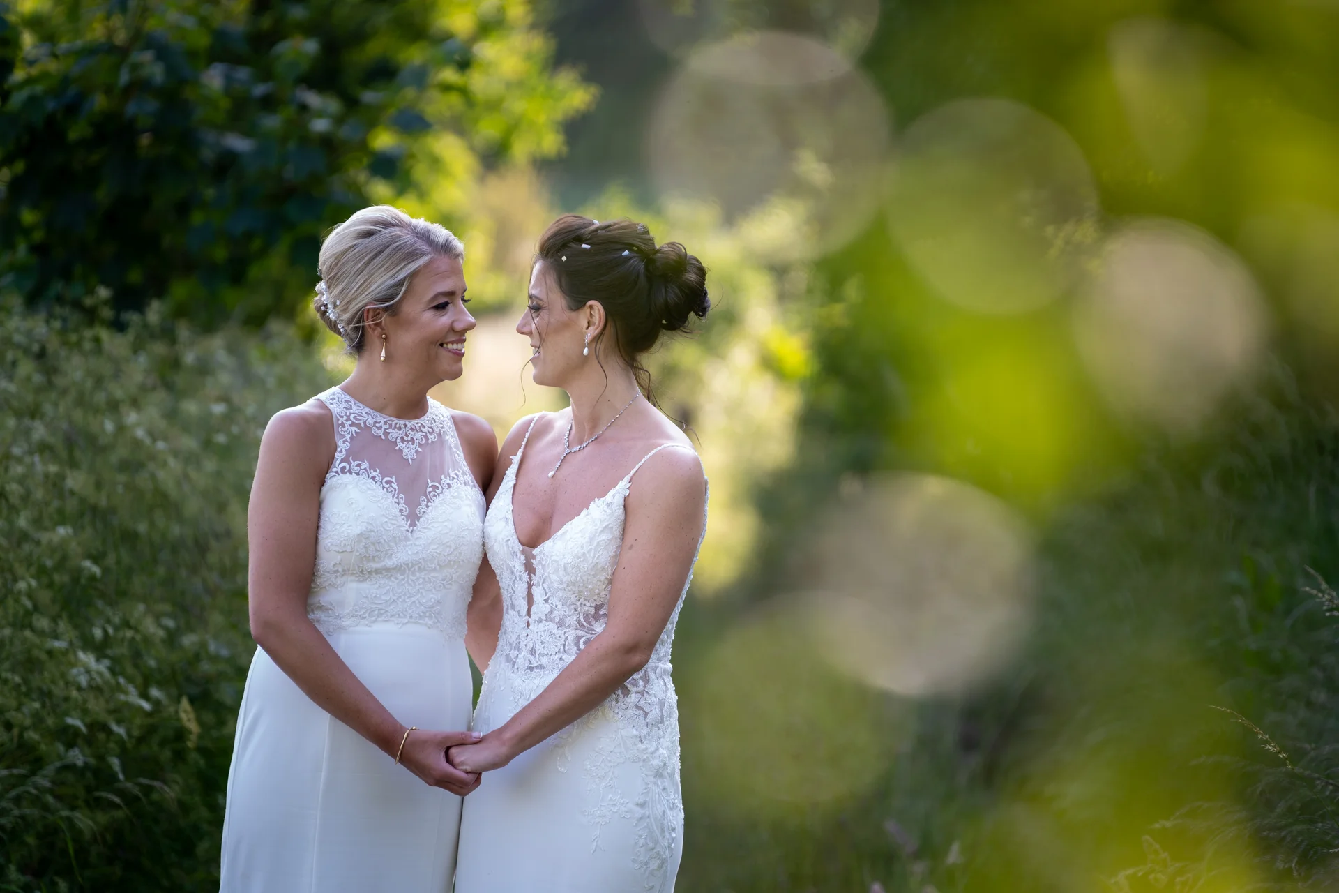 Two smiling brides in white wedding dresses stand outdoors by a lush green setting, holding hands and looking lovingly at each other through soft foliage bokeh.