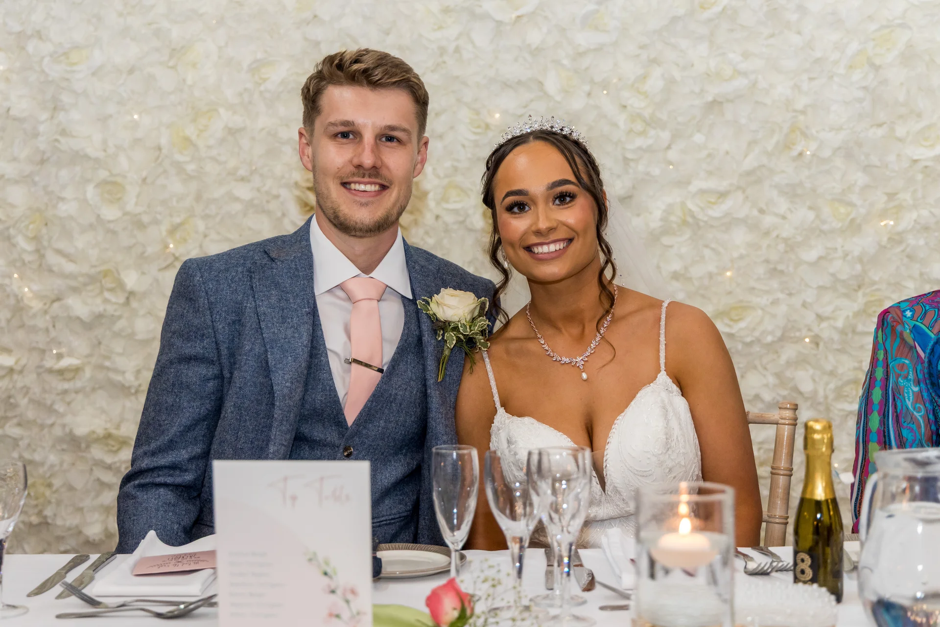 A smiling bride and groom sit together at the top table in front of a white floral wall, with the groom wearing a blue tweed-style suit and pink tie, and the bride wearing a white gown, crystal tiara, and statement necklace.