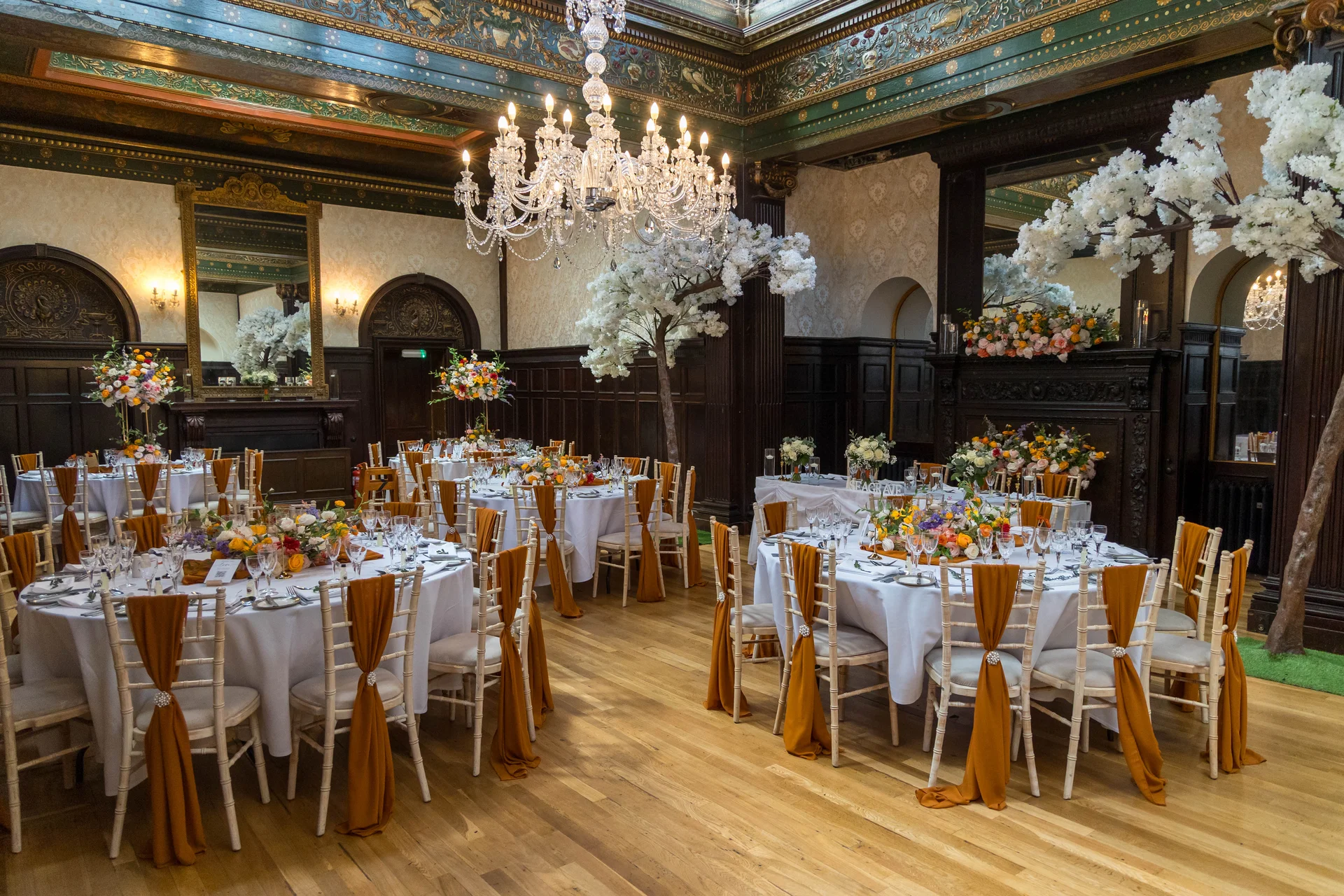 An elegant grand ballroom reception setup with round tables adorned with white linens, burnt orange chair sashes, and vibrant floral centrepieces beneath a striking crystal chandelier.