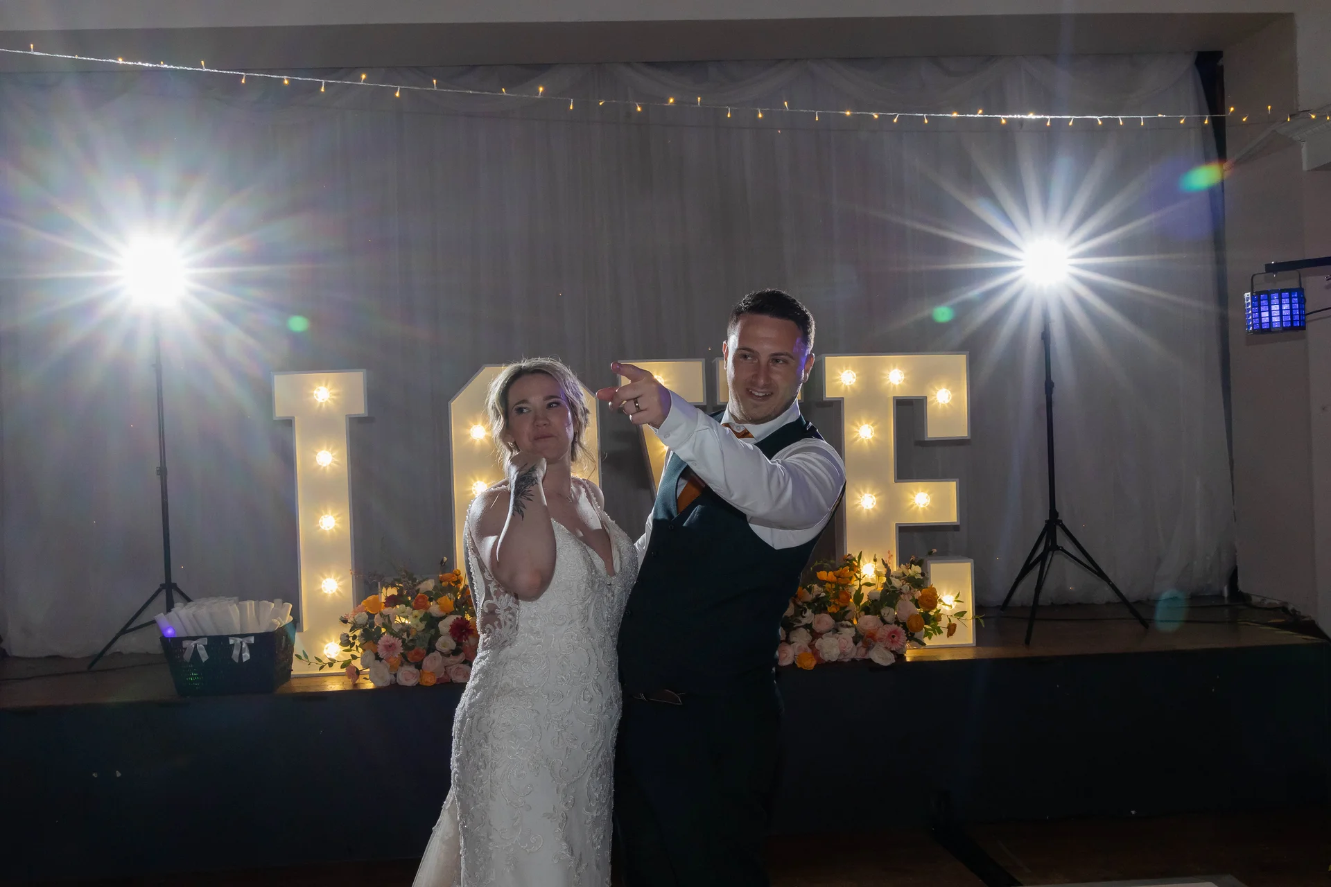 A newlywed couple poses playfully in front of large illuminated love letters during their evening reception, framed by bright stage lights and dramatic lens flare.