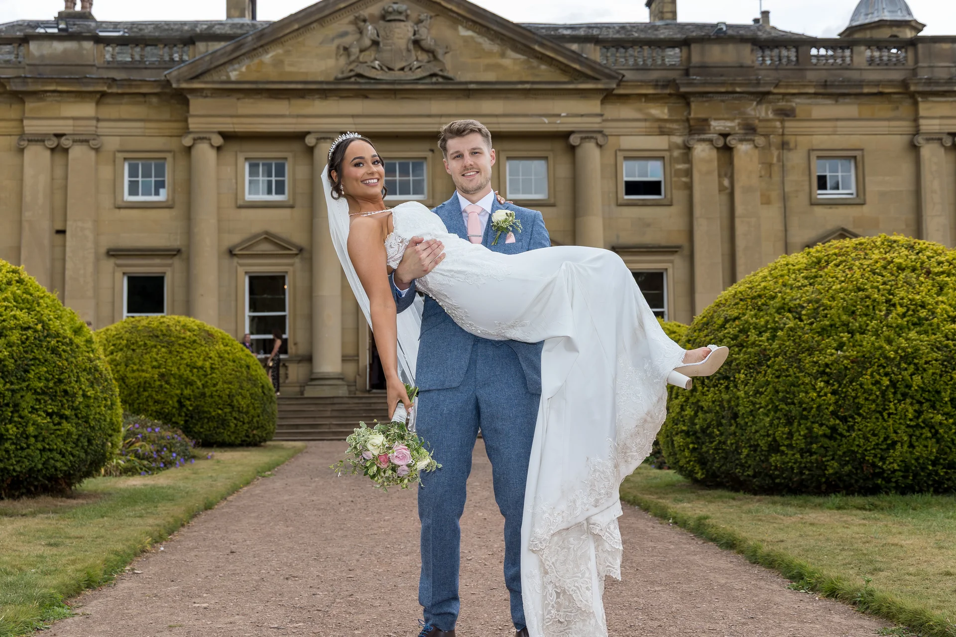 A groom proudly lifts his smiling bride in his arms in front of a grand country house with manicured topiary bushes lining the gravel path.
