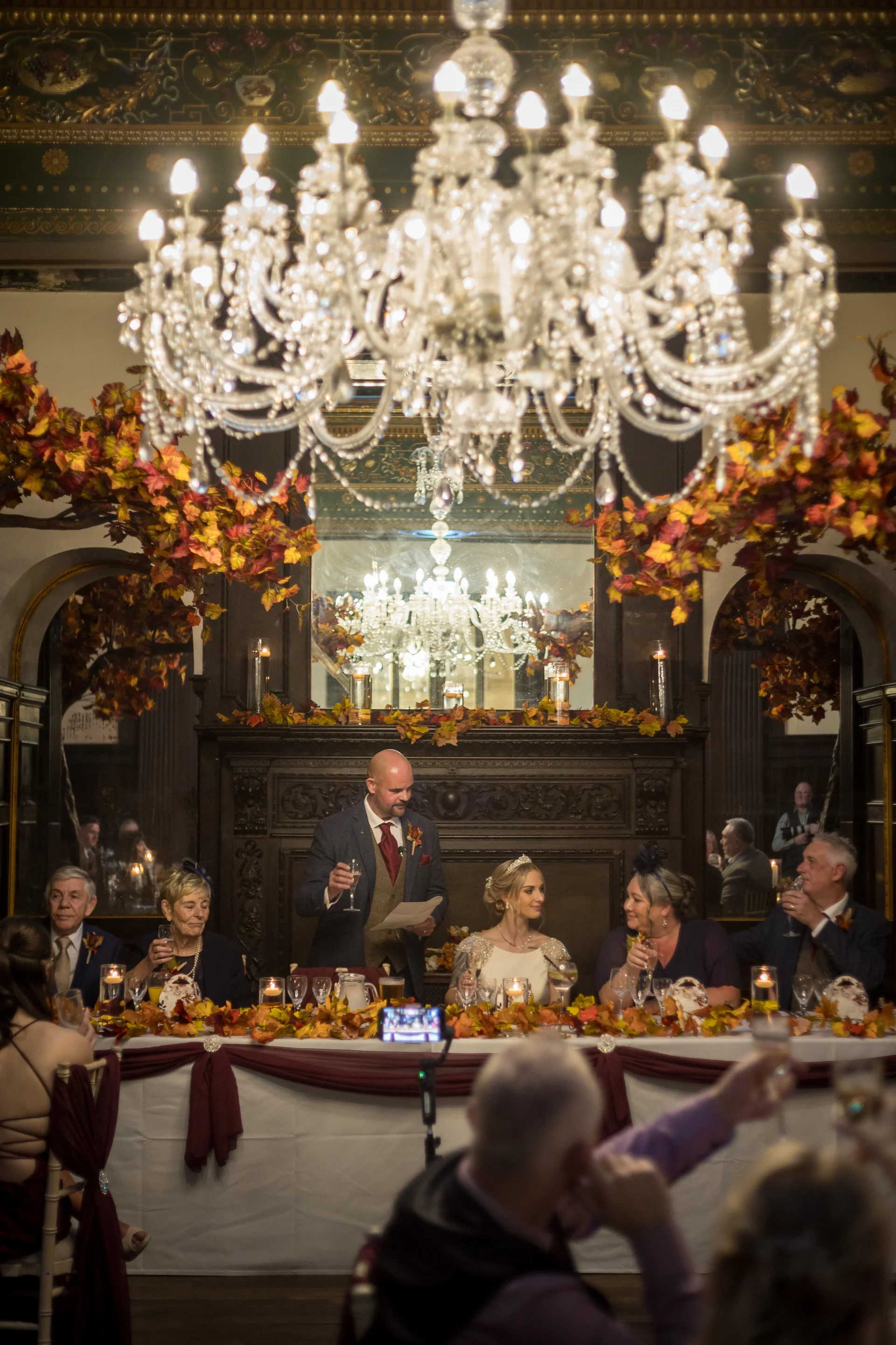 The groom delivers a speech at the top table during an elegant autumn wedding reception, illuminated by a grand crystal chandelier and warm candlelight.