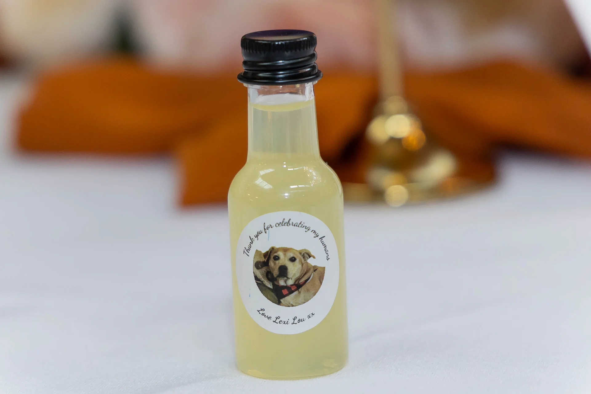 A miniature glass bottle wedding favour filled with a yellow drink features a custom sticker of a dog with text reading thank you for celebrating my humans.