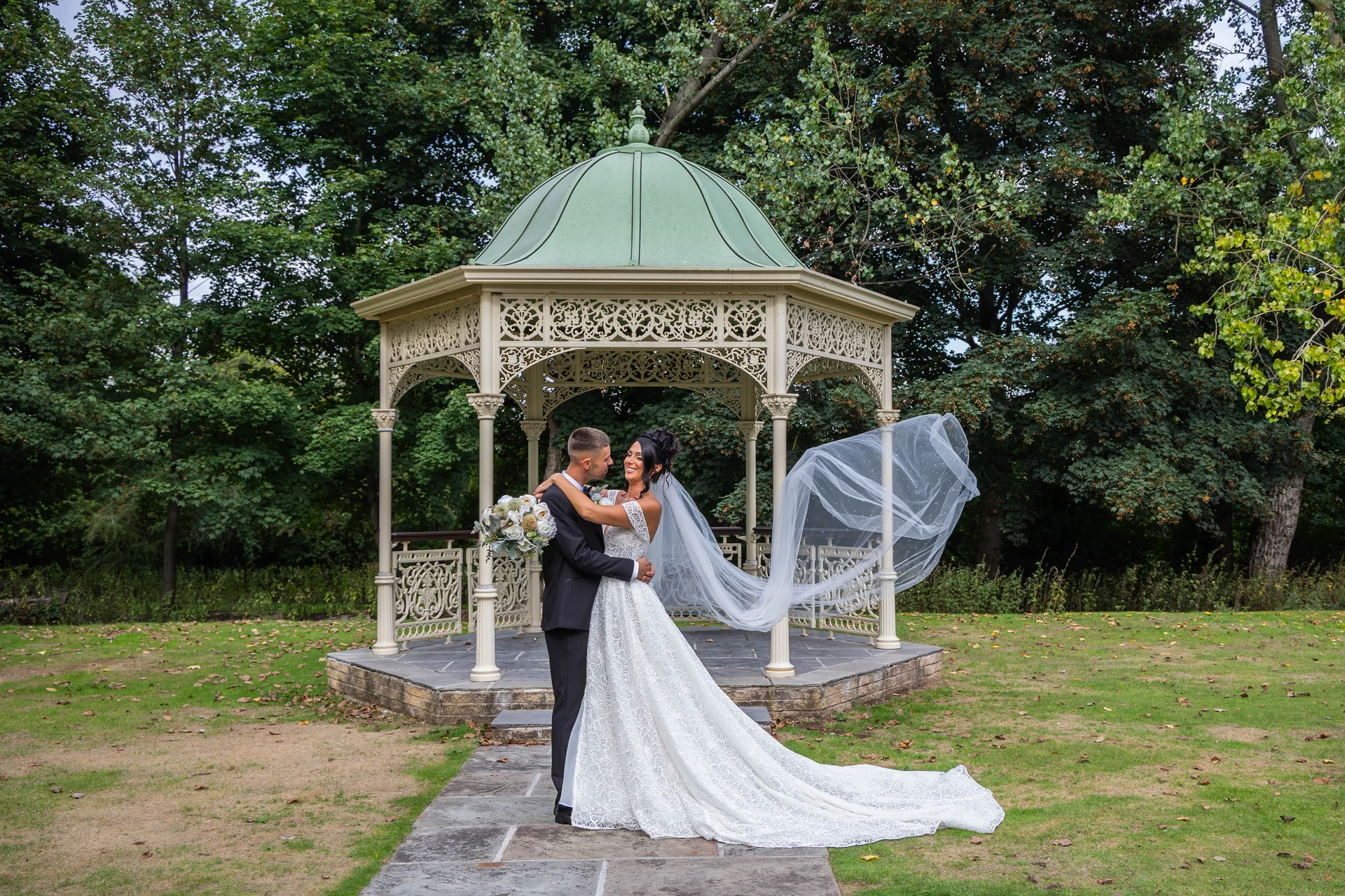 A newlywed couple embraces lovingly in front of an ornate garden bandstand, with the bride's cathedral veil dramatically catching the wind.