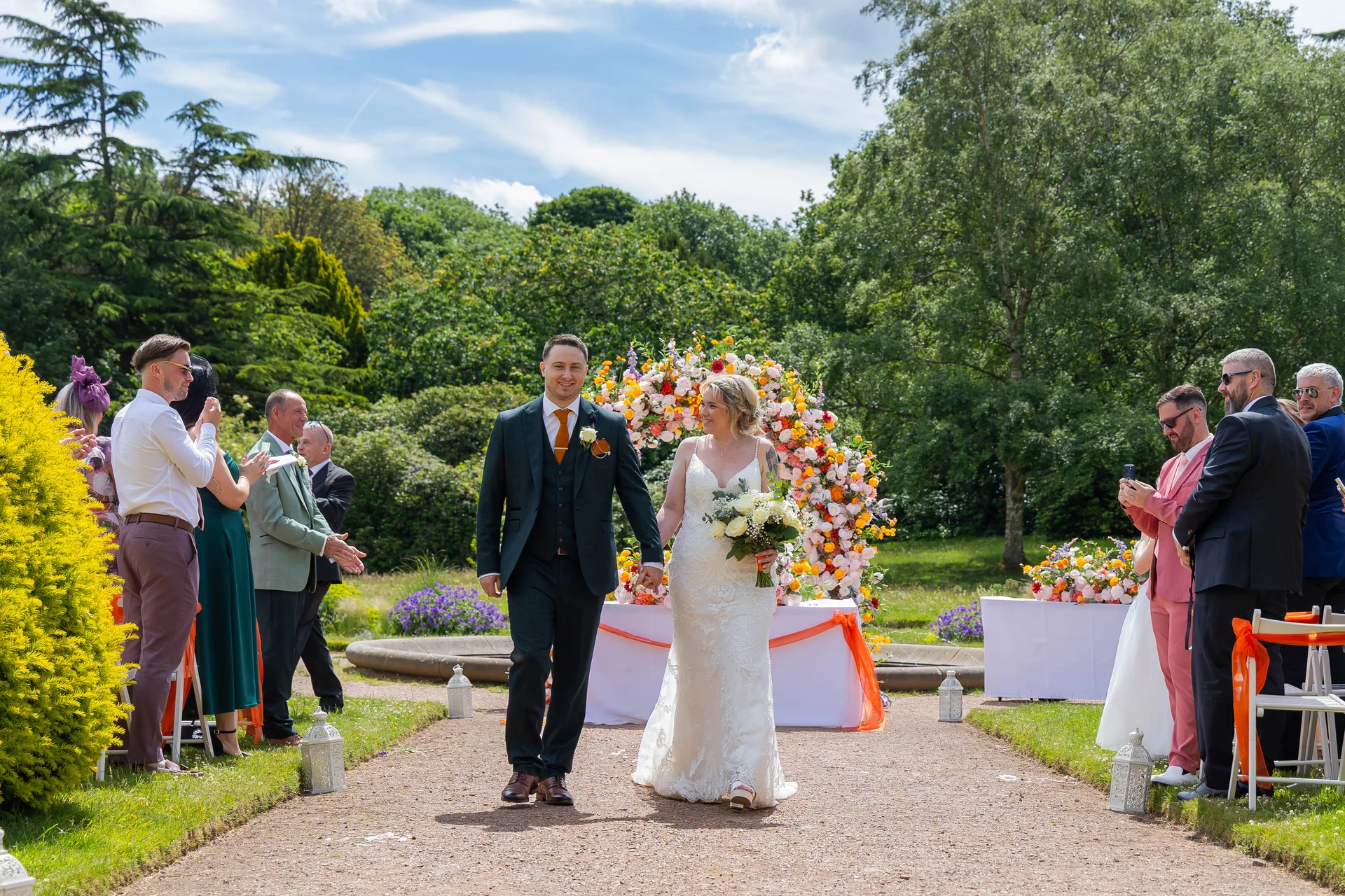 A newlywed couple happily walks hand in hand down the garden aisle after their outdoor ceremony, flanked by cheering guests and a vibrant floral arch.