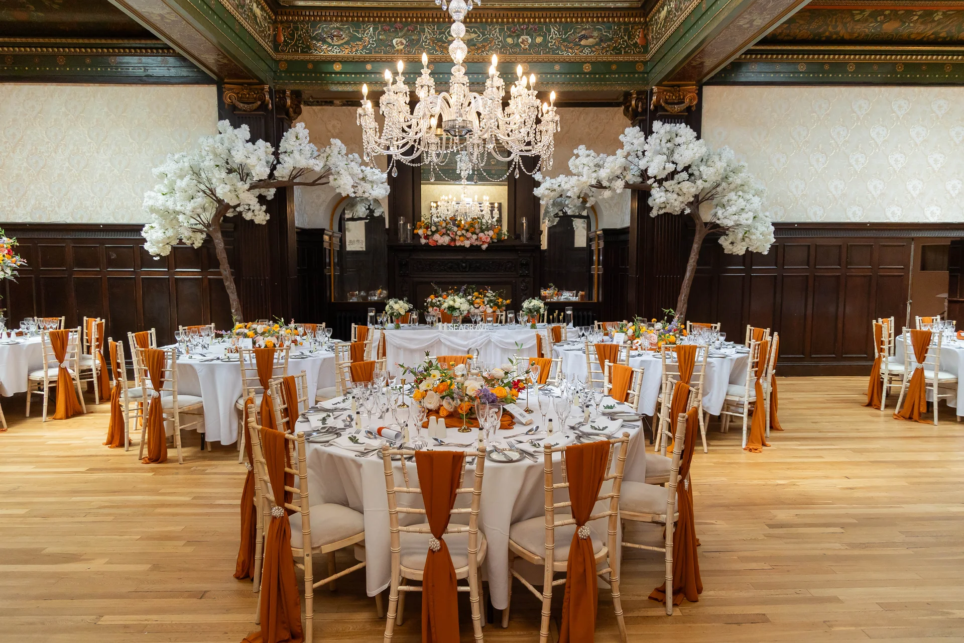 A grand stately home ballroom is elegantly set up for a wedding breakfast with round tables featuring white linens, burnt orange sashes, and vibrant floral centerpieces beneath a magnificent crystal chandelier.
