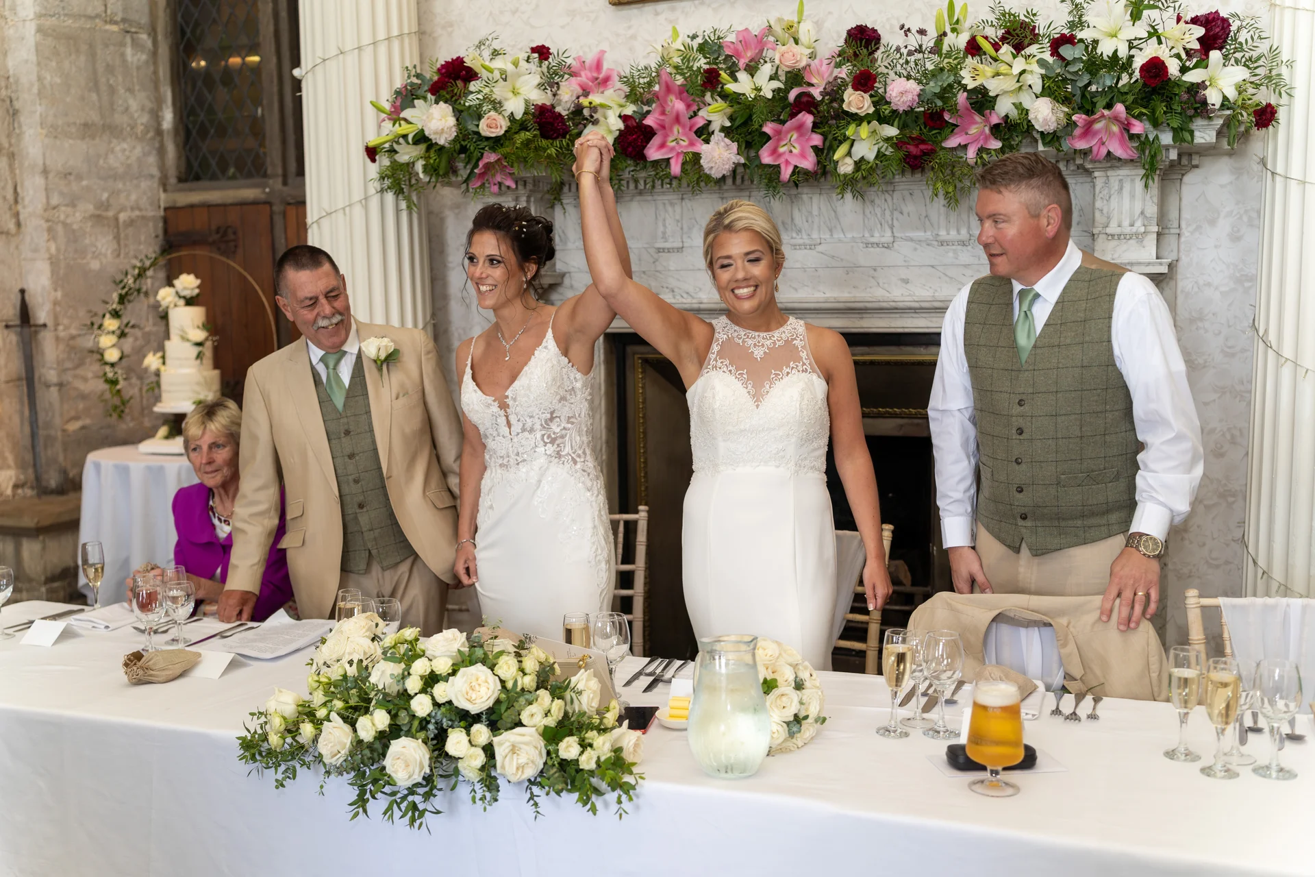 Two smiling brides hold hands in the air during the wedding breakfast, flanked by family members in front of a grand floral mantelpiece.