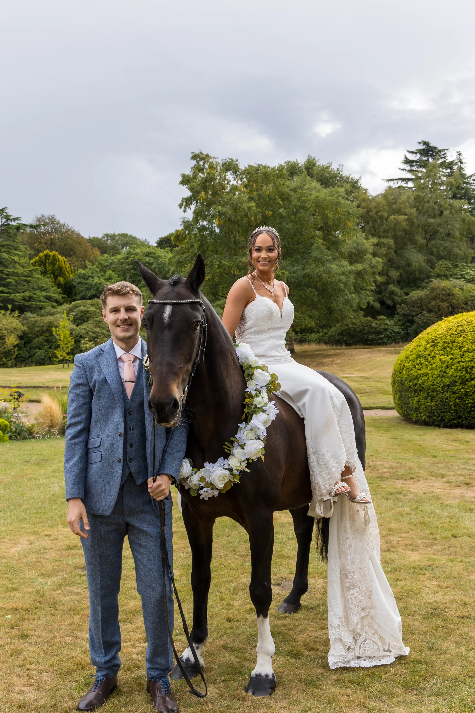 A smiling groom in a blue tweed suit stands beside a dark horse while his bride in a white lace gown and tiara sits gracefully on its back in a scenic garden.