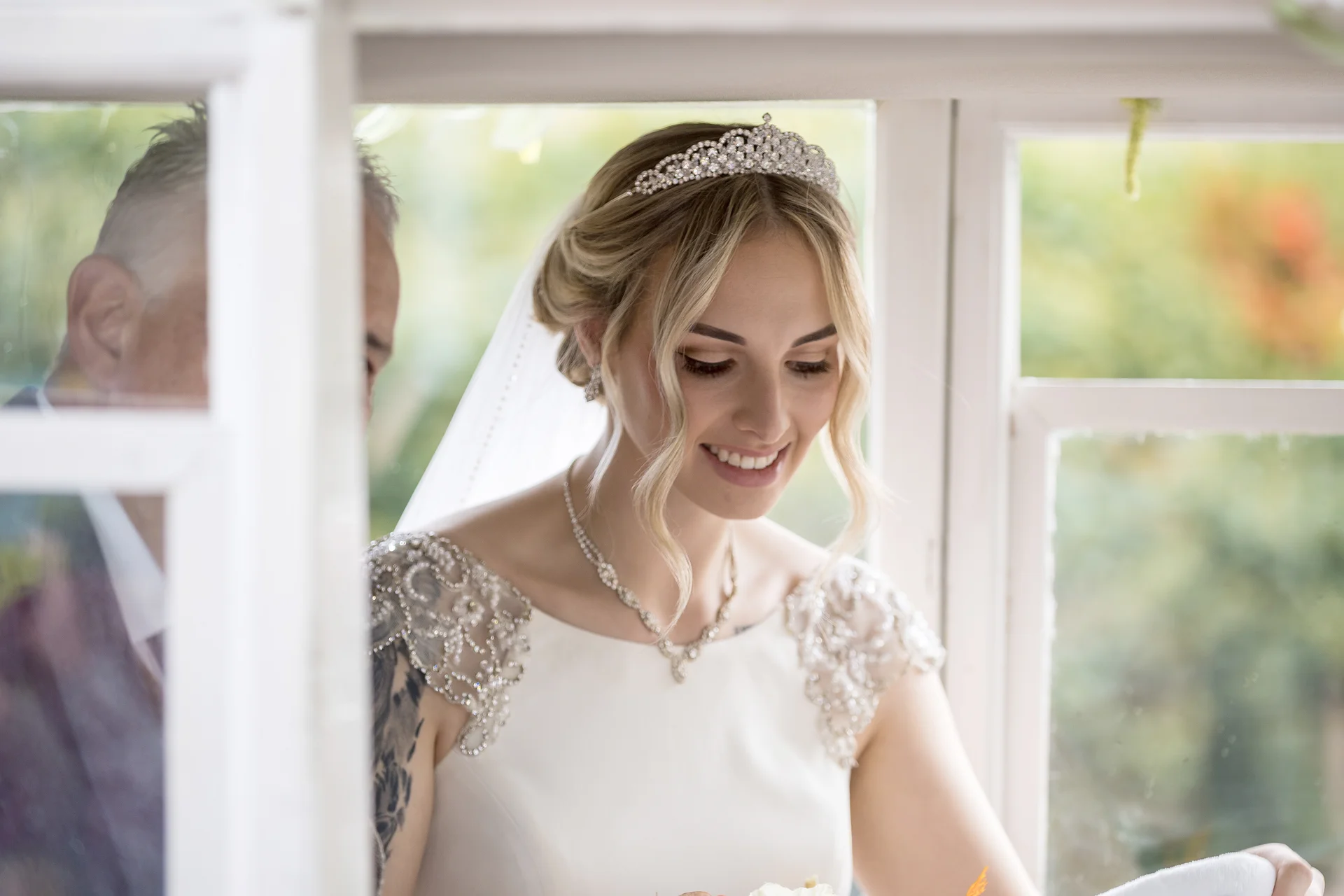A smiling bride wearing a crystal tiara and beaded gown stands next to her father framed by a doorway before the ceremony.