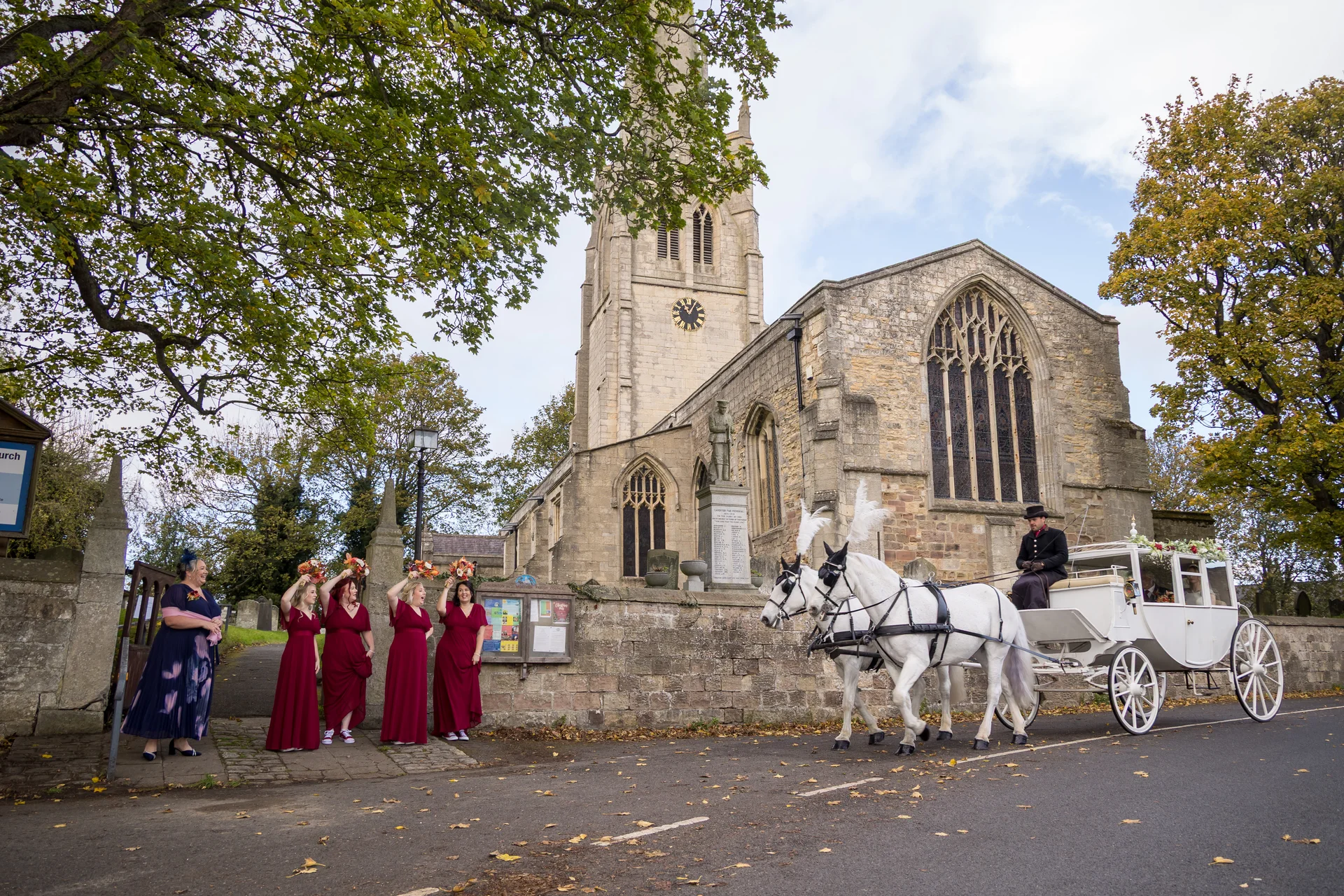 Bridesmaids in red dresses wave as a magnificent white horse-drawn carriage arrives outside an historic stone church for the wedding ceremony.