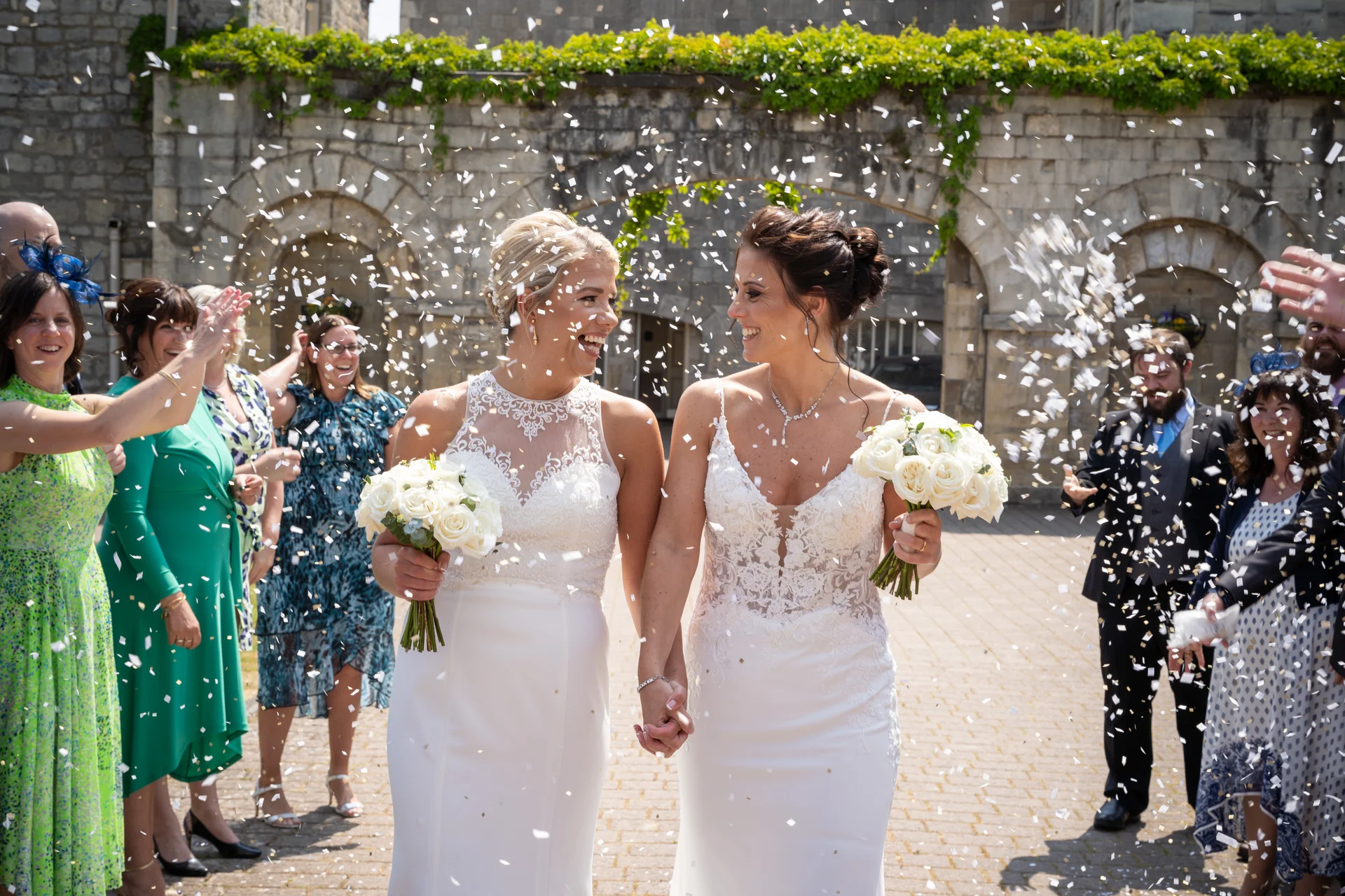 Two smiling brides holding hands and bouquets as guests shower them with white confetti outside a historic stone building.