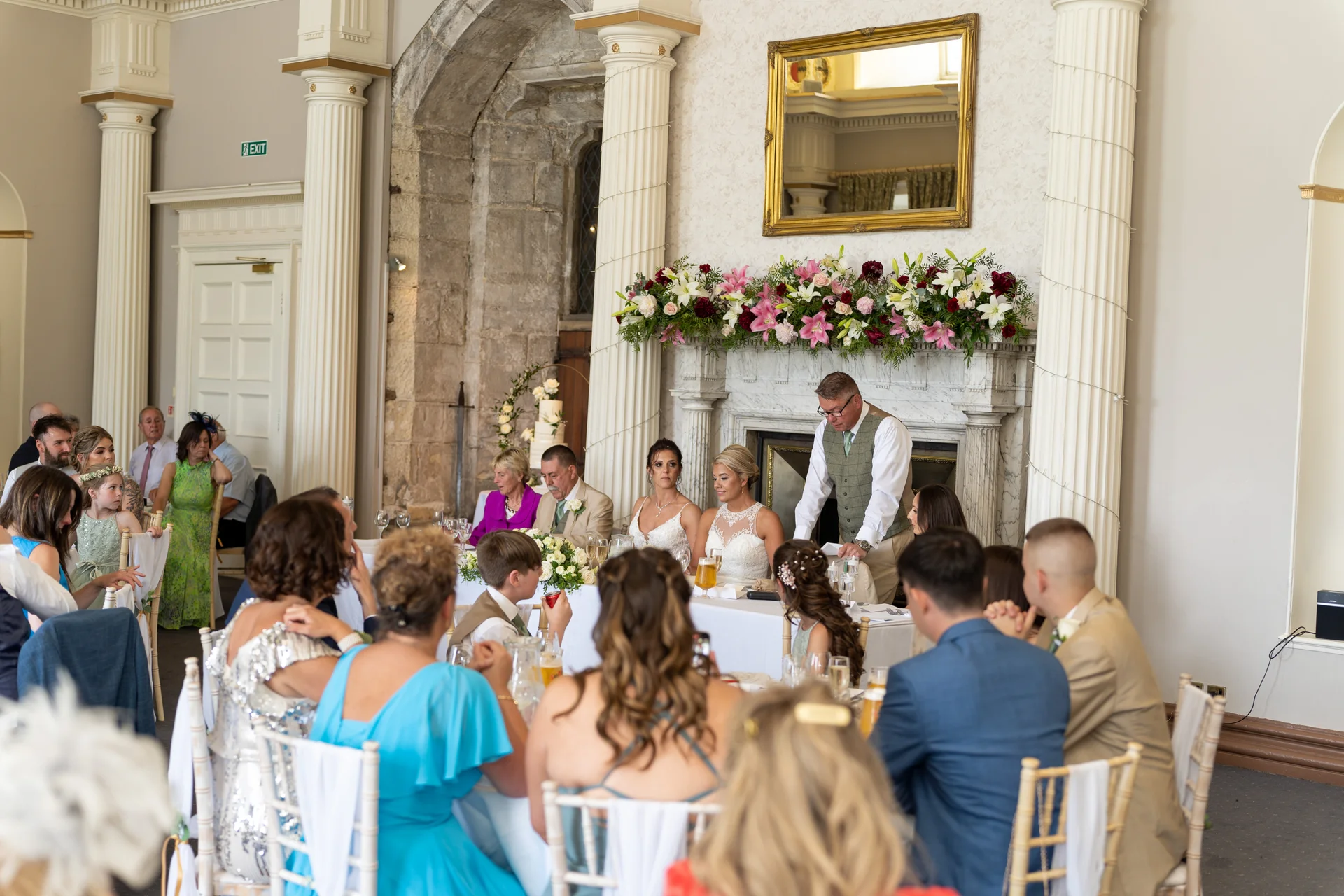 A wedding breakfast takes place inside a grand historic country house hall where a speaker delivers a speech at the top table.