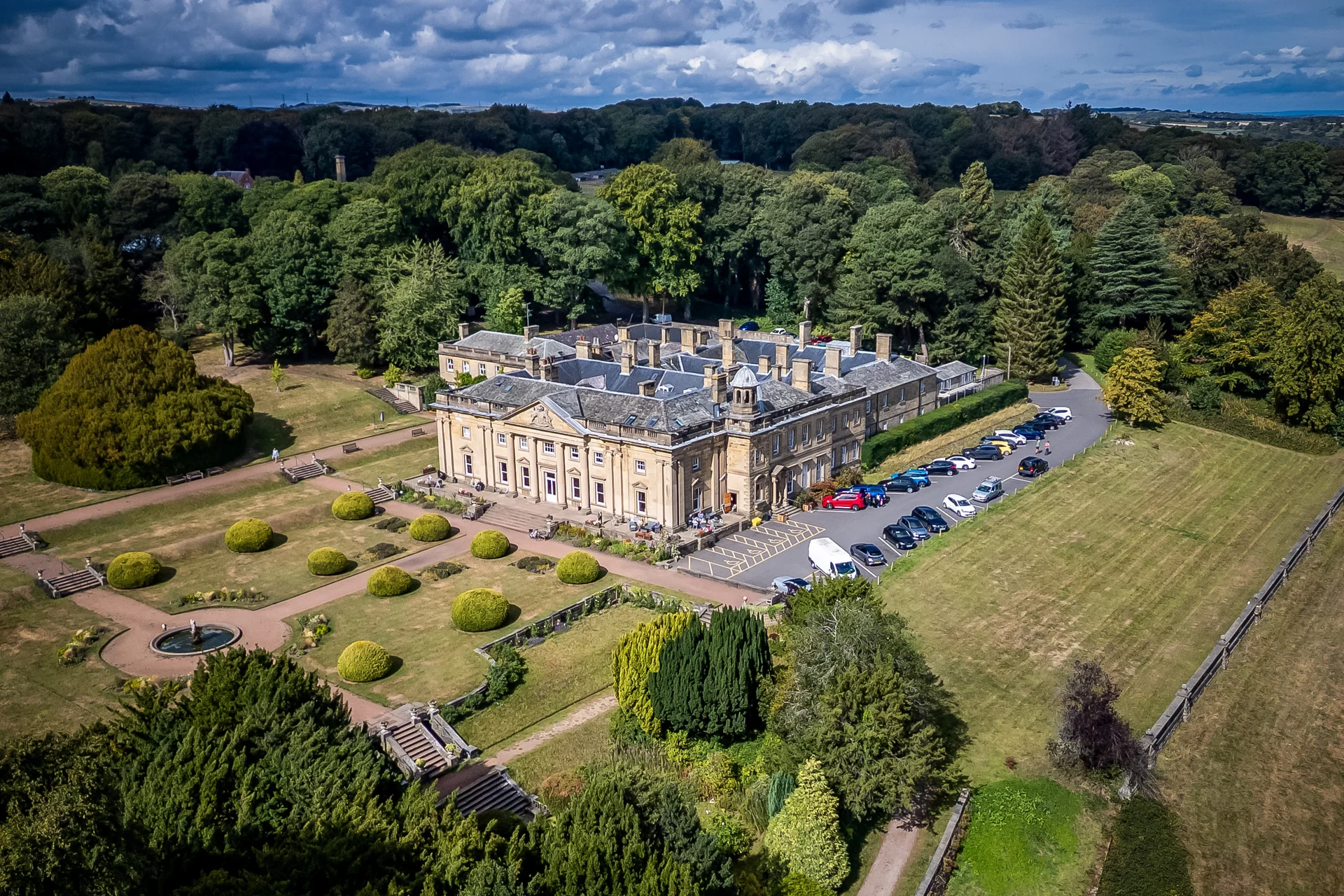 An aerial drone view captures a magnificent country house wedding venue surrounded by lush manicured gardens and dense woodland under a dramatic sky.
